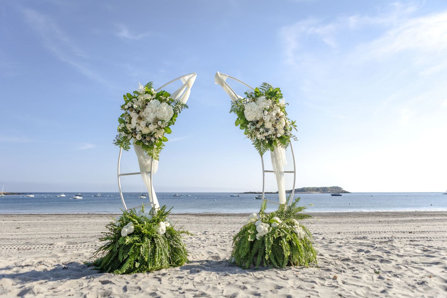 Two elegant floral arch arrangements with white flowers and greenery on a sandy beach with ocean views.