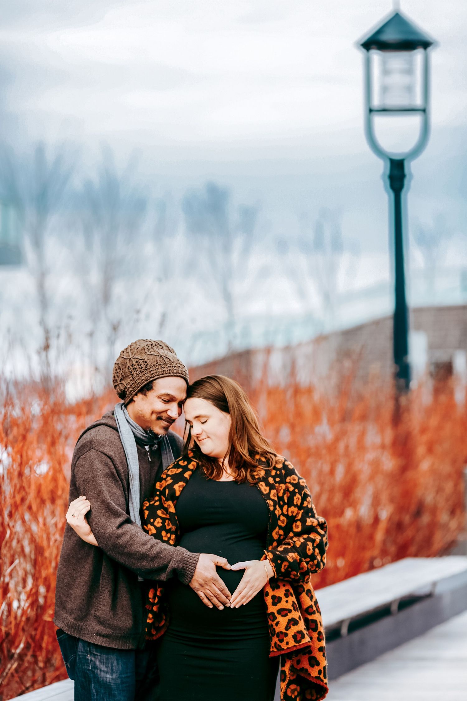 Couple making heart shape on pregnant belly outdoors in winter near red bushes and street lamp.
