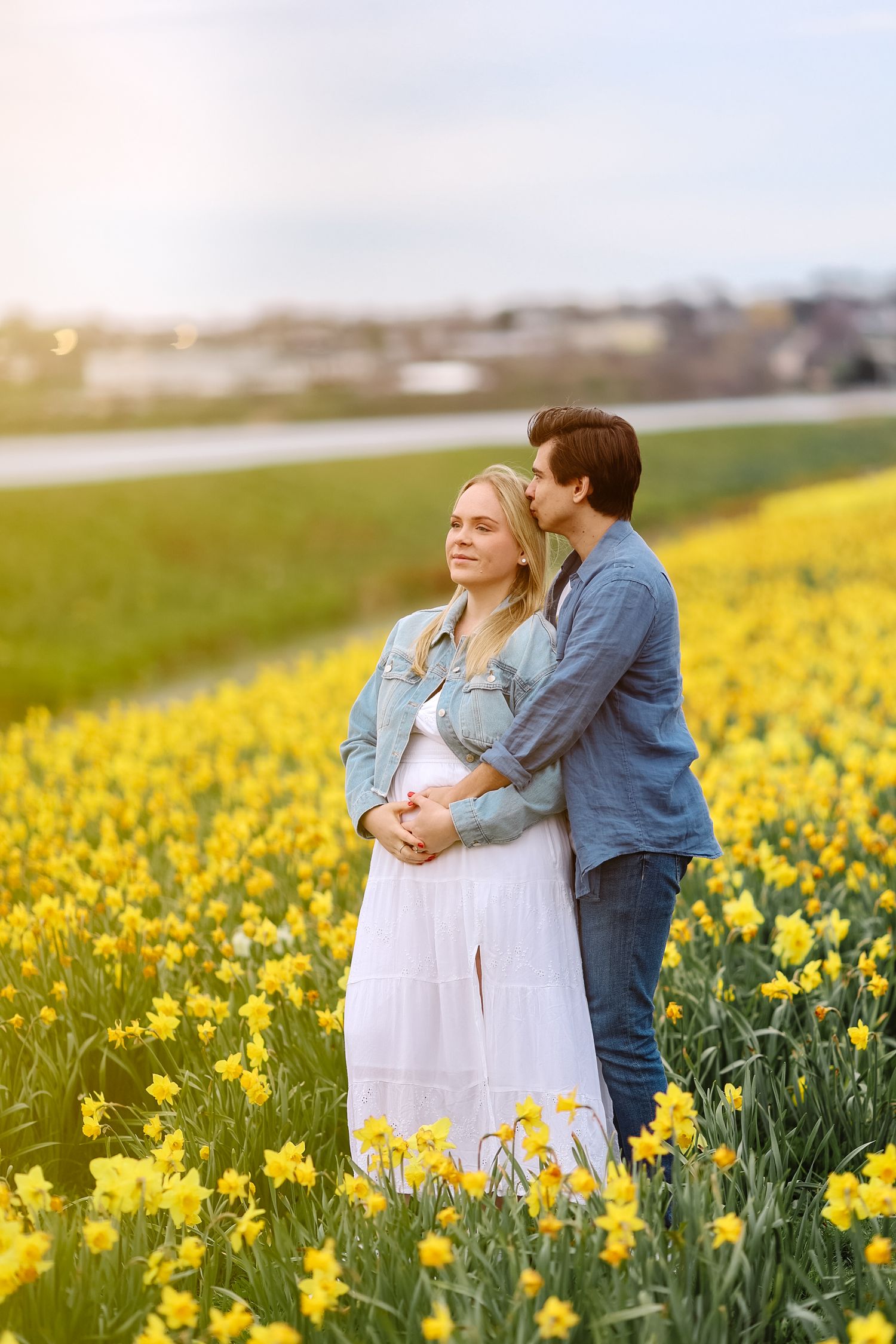 Man kissing pregnant woman on cheek while embracing her from behind in a sunny yellow daffodil field.