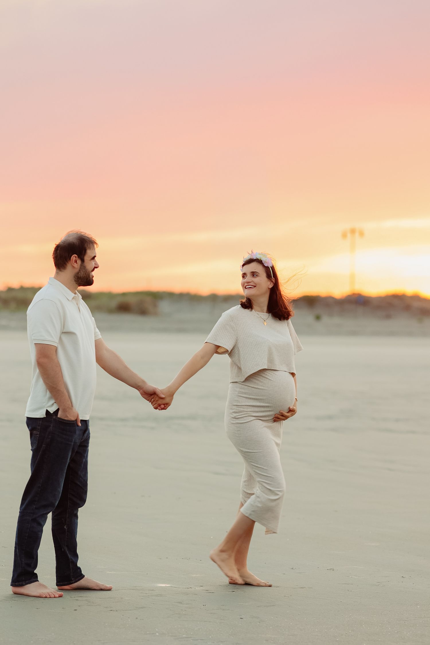 Pregnant couple holding hands on sandy beach at sunset, woman in white outfit smiling at her partner.