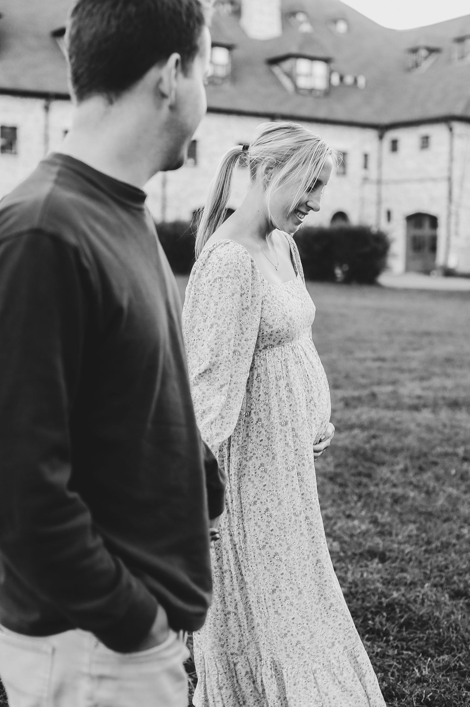 Black and white photo of pregnant woman in floral dress smiling at baby bump beside partner outdoors.