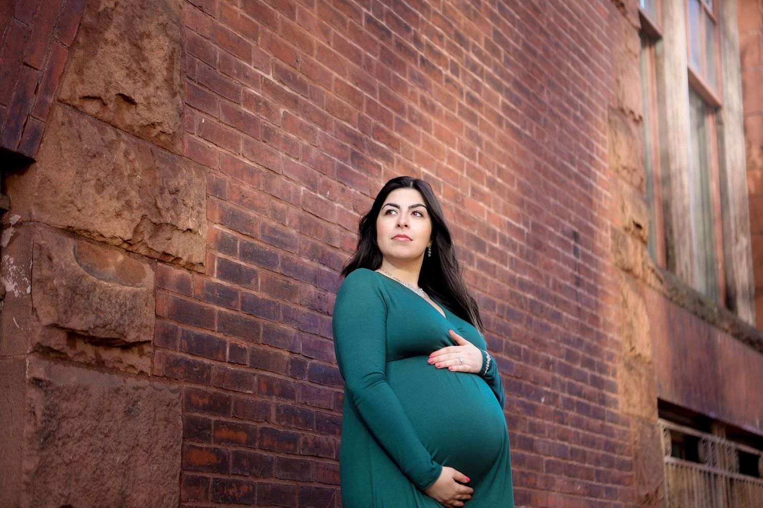 Pregnant woman in green dress standing against rustic brick wall cradling her baby bump outdoors.