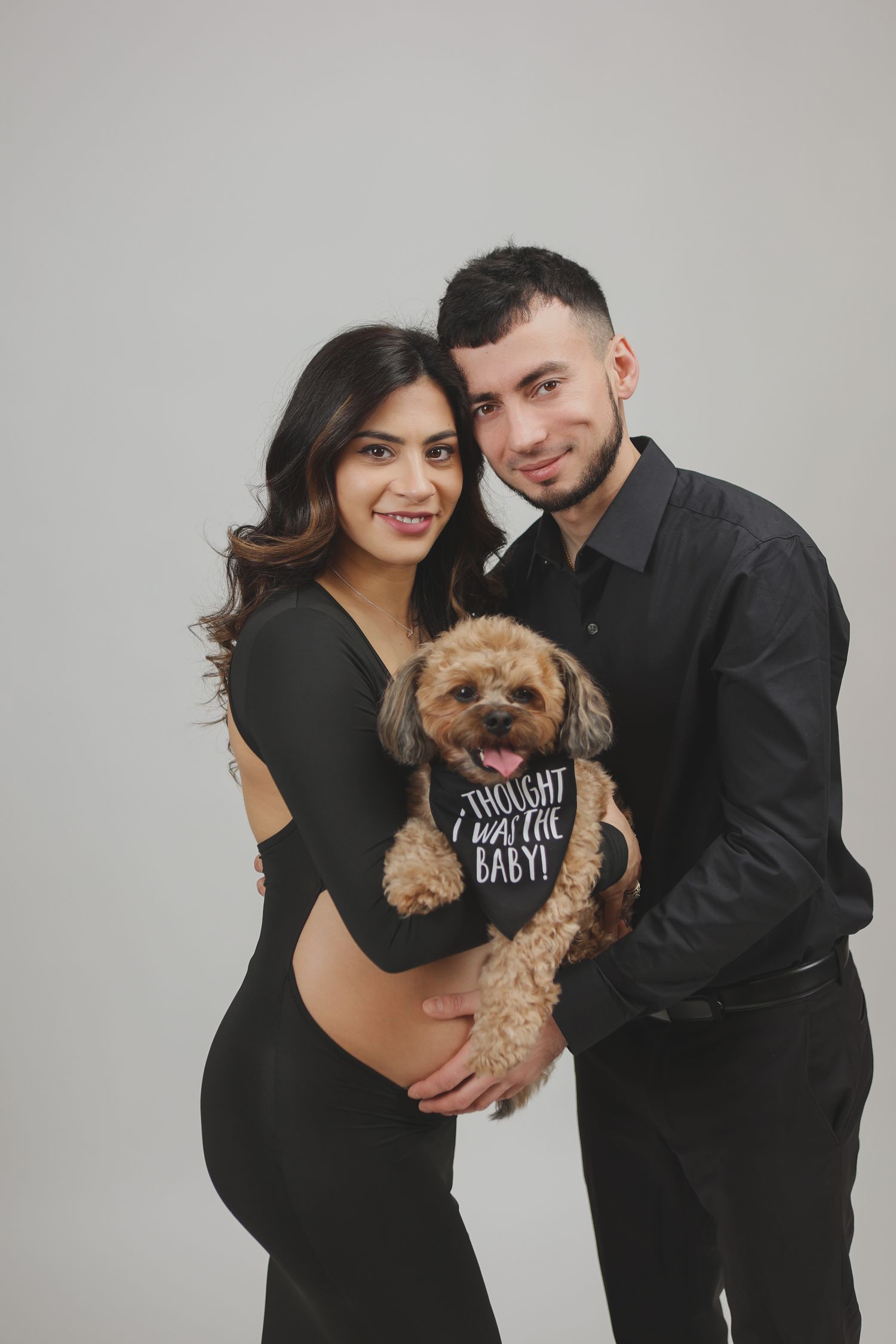 Smiling expecting couple in black outfits holding golden doodle with I thought I was the baby bandana.
