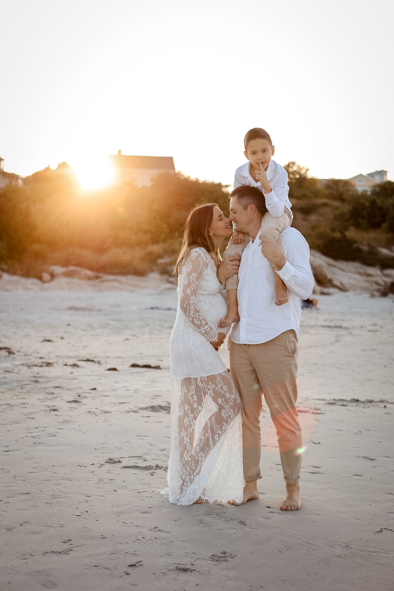 Family with boy on shoulders kissing at golden sunset beach.