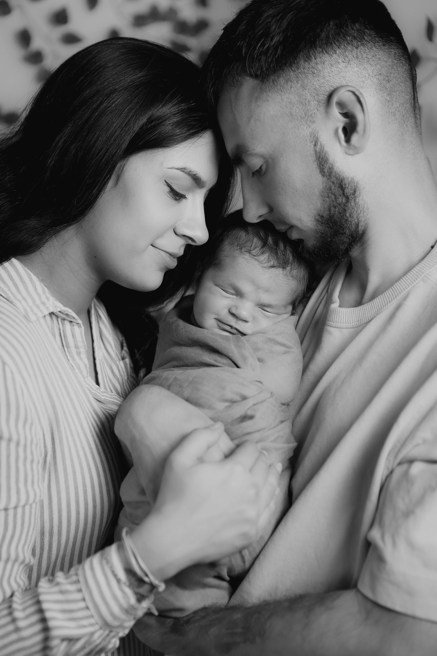 Black and white portrait of parents touching foreheads while tenderly holding sleeping newborn baby.