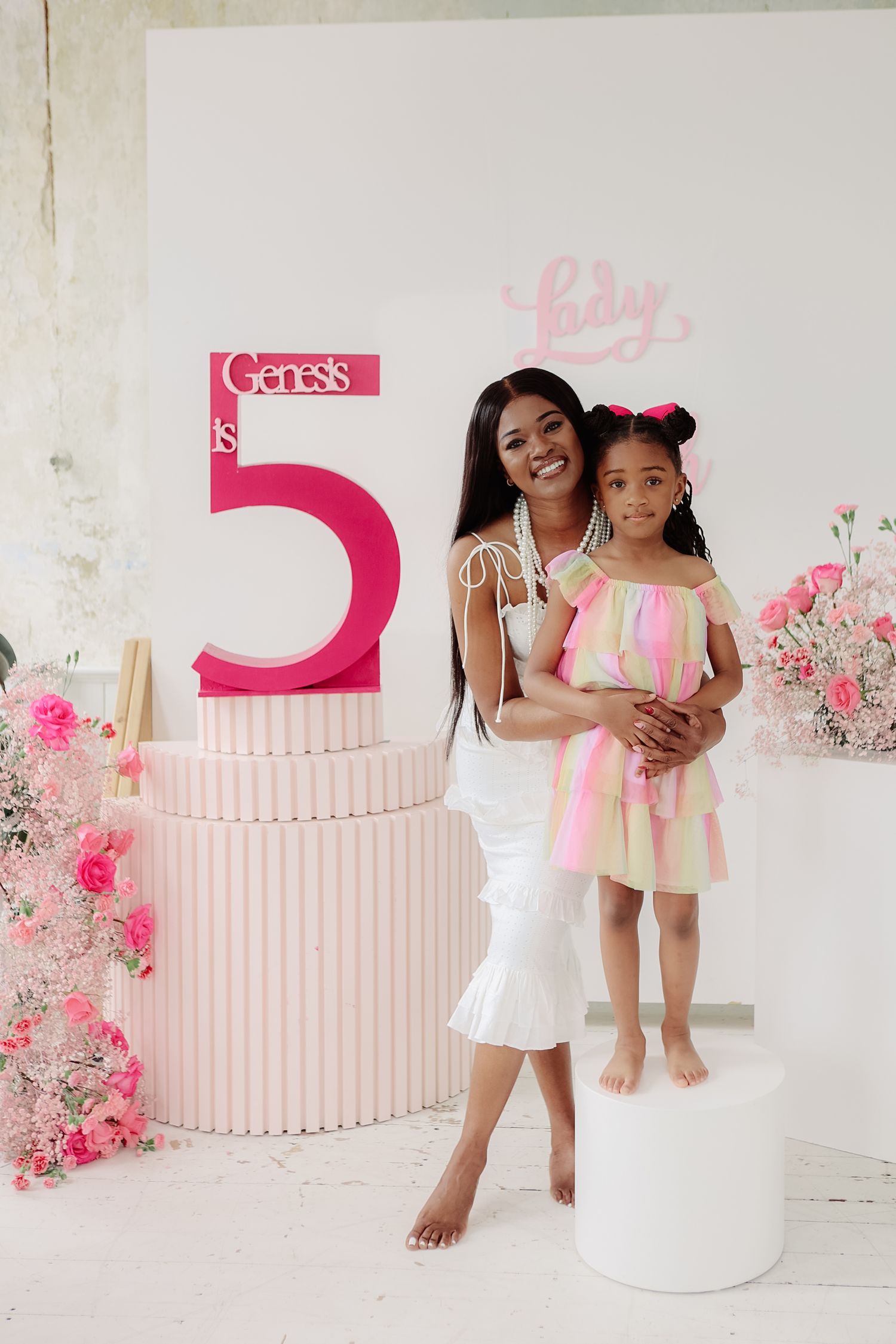 Woman in white dress holds young girl at Genesis's 5th birthday party with pink floral decorations.