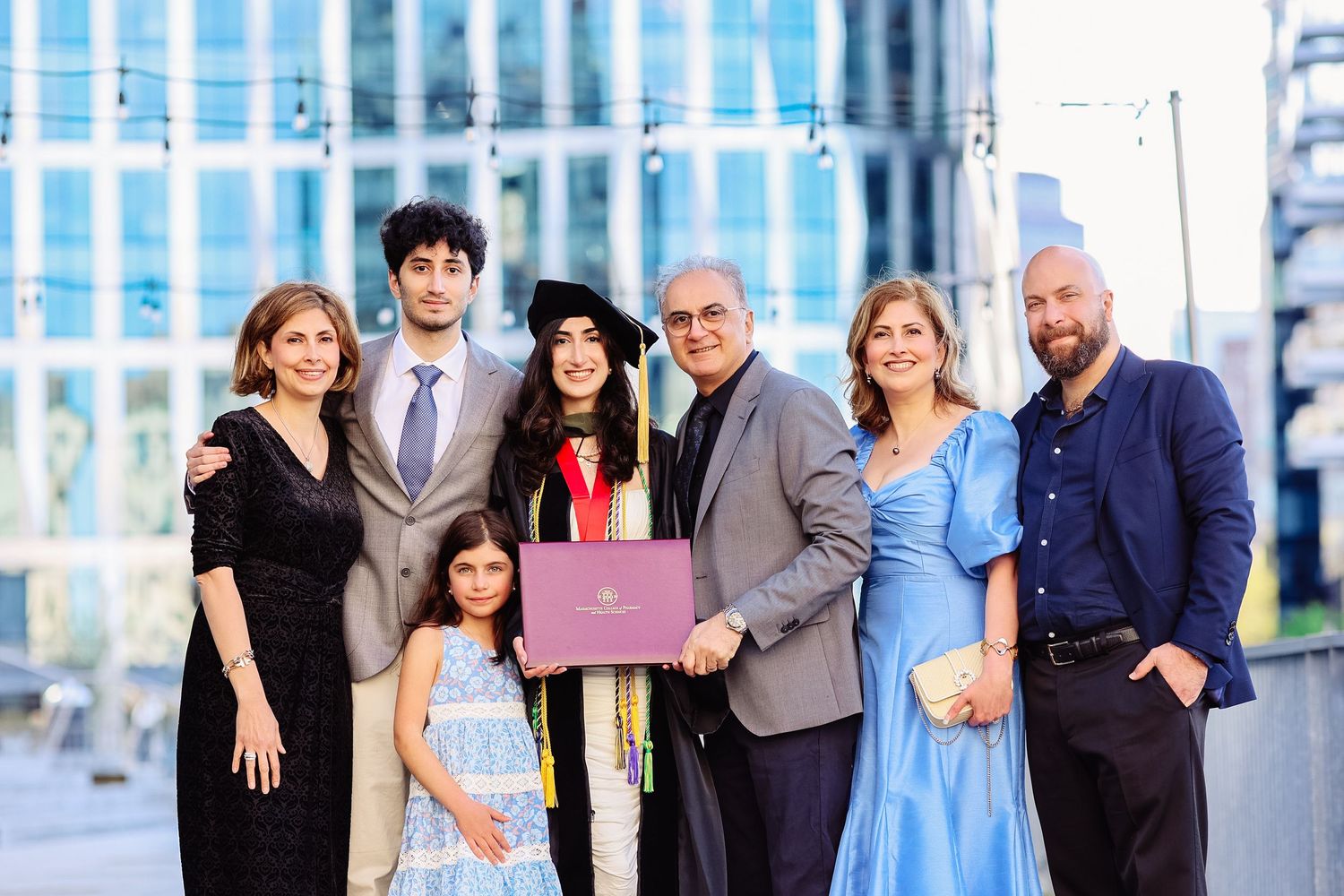 Family celebrating graduation outdoors, graduate holding diploma in cap and gown with seven relatives.