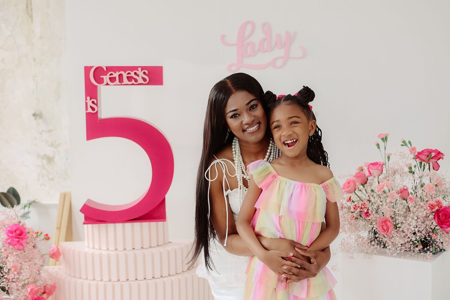 Woman in white dress hugging smiling girl in colorful dress at Genesis 5th birthday party.