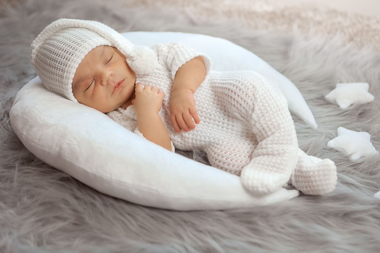 Peaceful newborn baby in white knit bunny outfit sleeps on crescent moon pillow on fur rug.