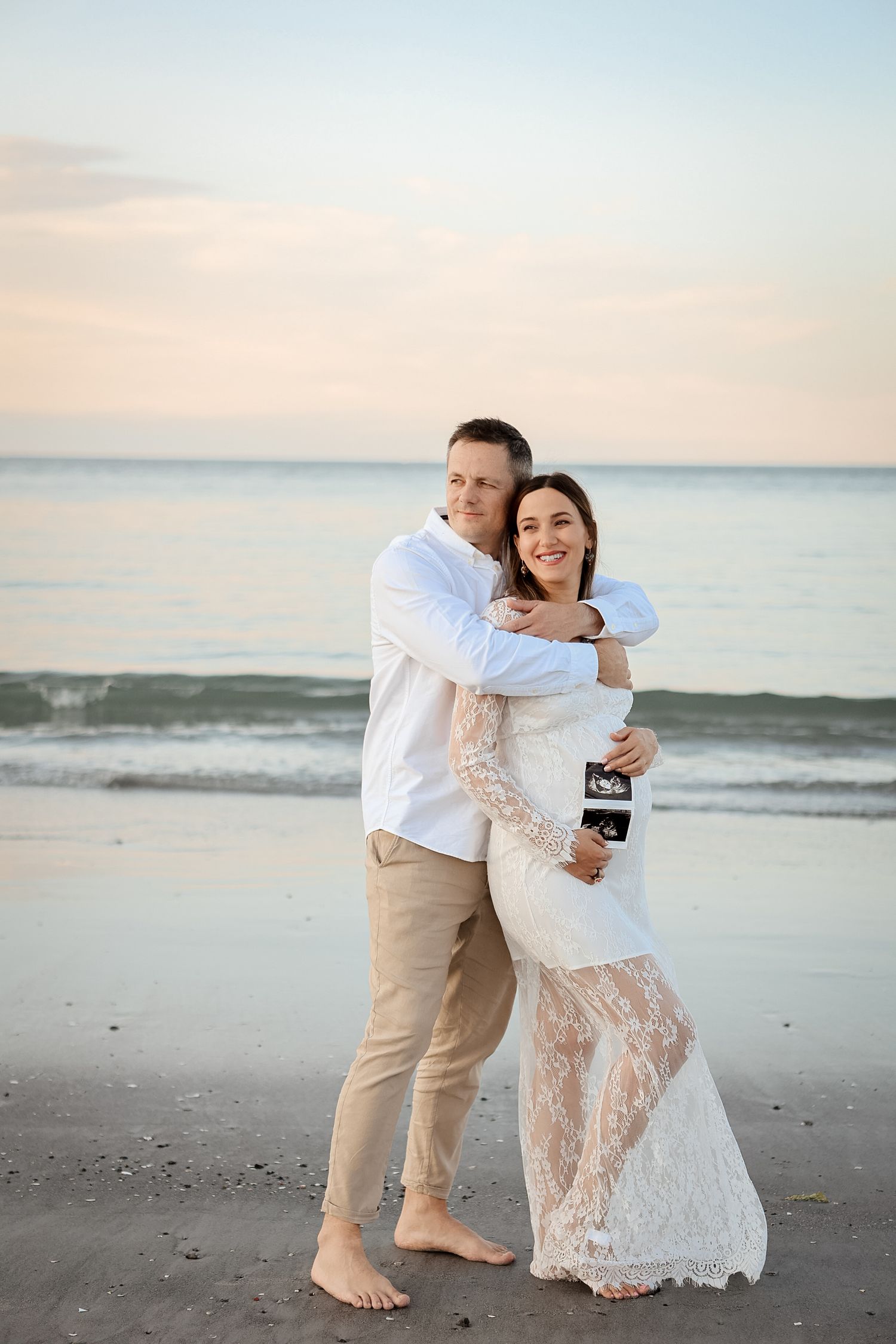 Couple embracing on beach at sunset, pregnant woman in lace dress holding ultrasound photo.