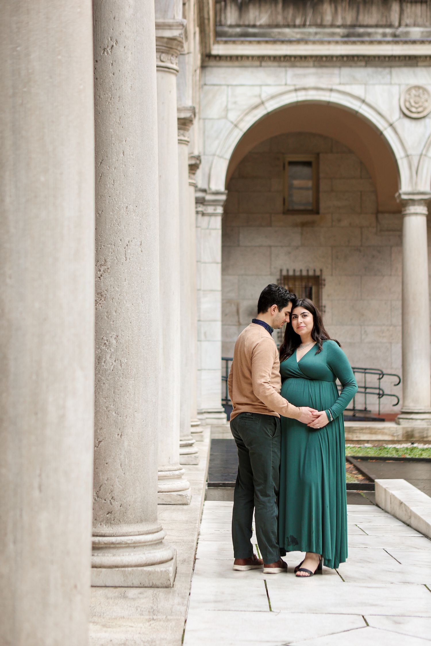 Expecting couple in green and tan outfits sharing tender moment between elegant stone columns.