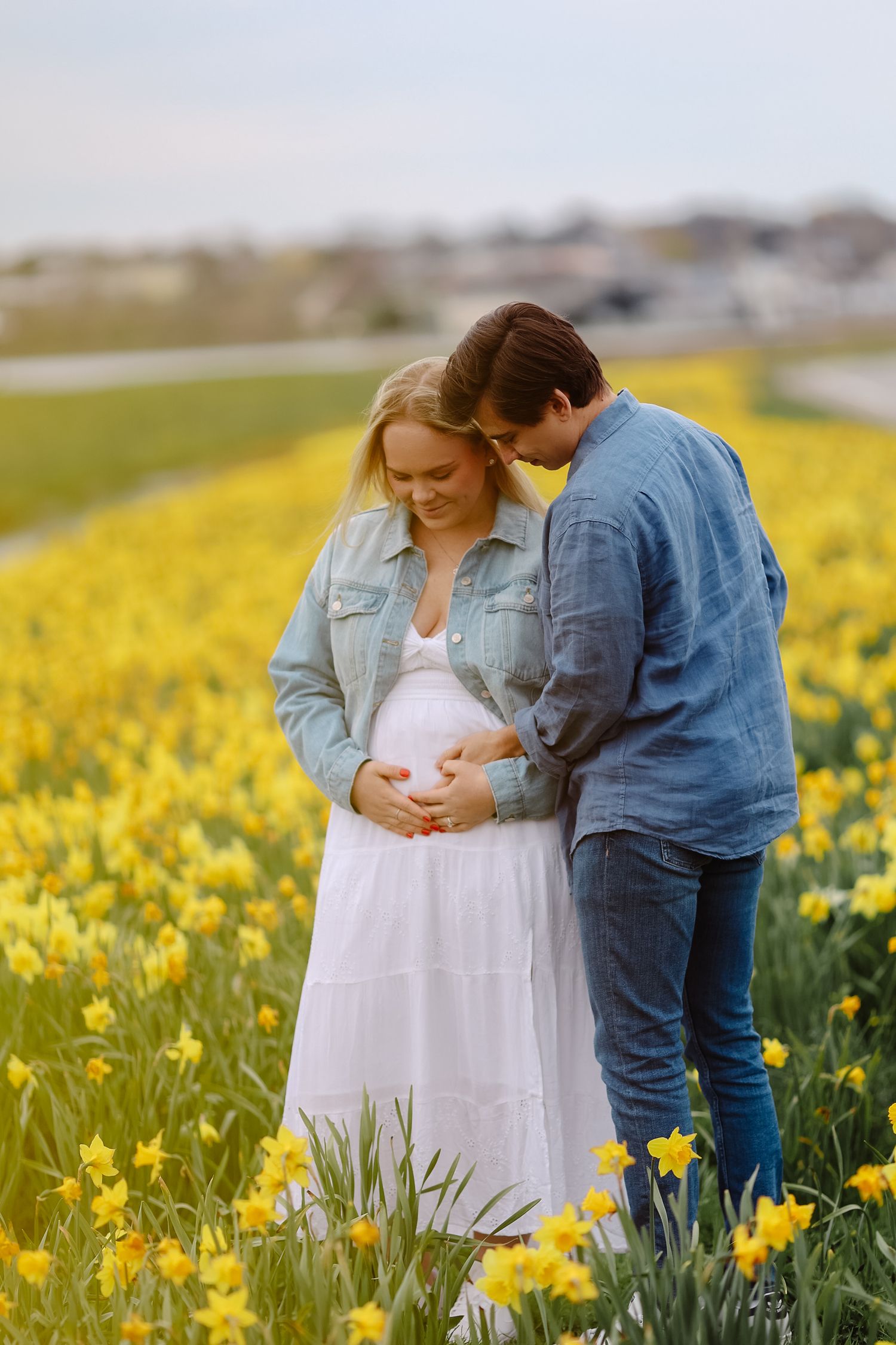 Couple cradling pregnant belly together in a bright yellow daffodil field, sharing a tender moment.