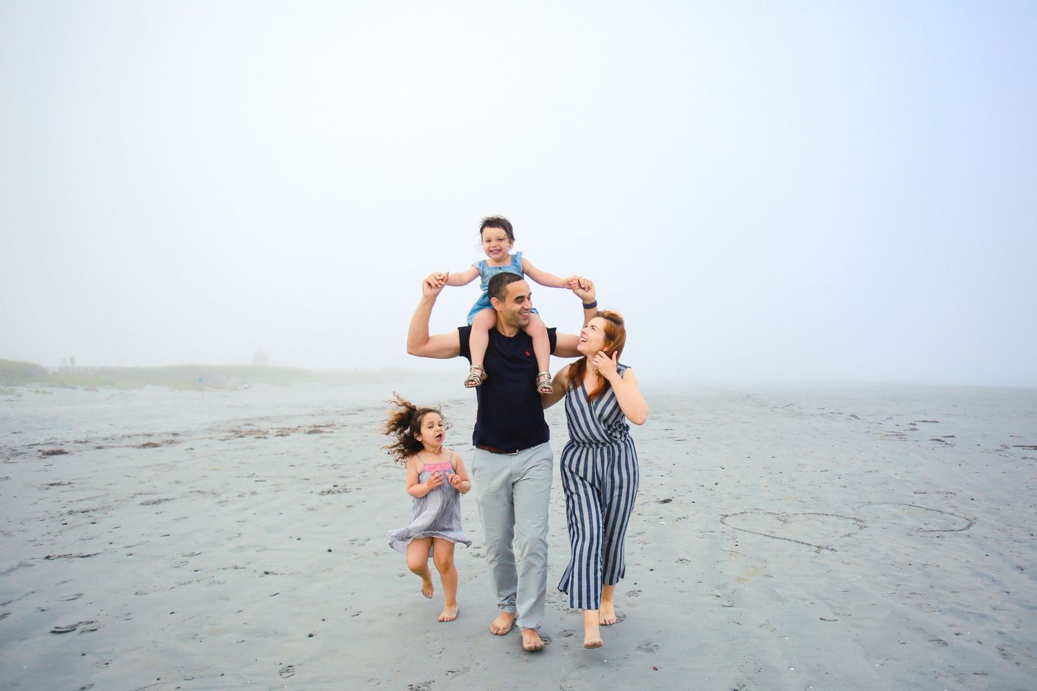 Family of four enjoying a foggy beach day, father with son on shoulders and daughter dancing.