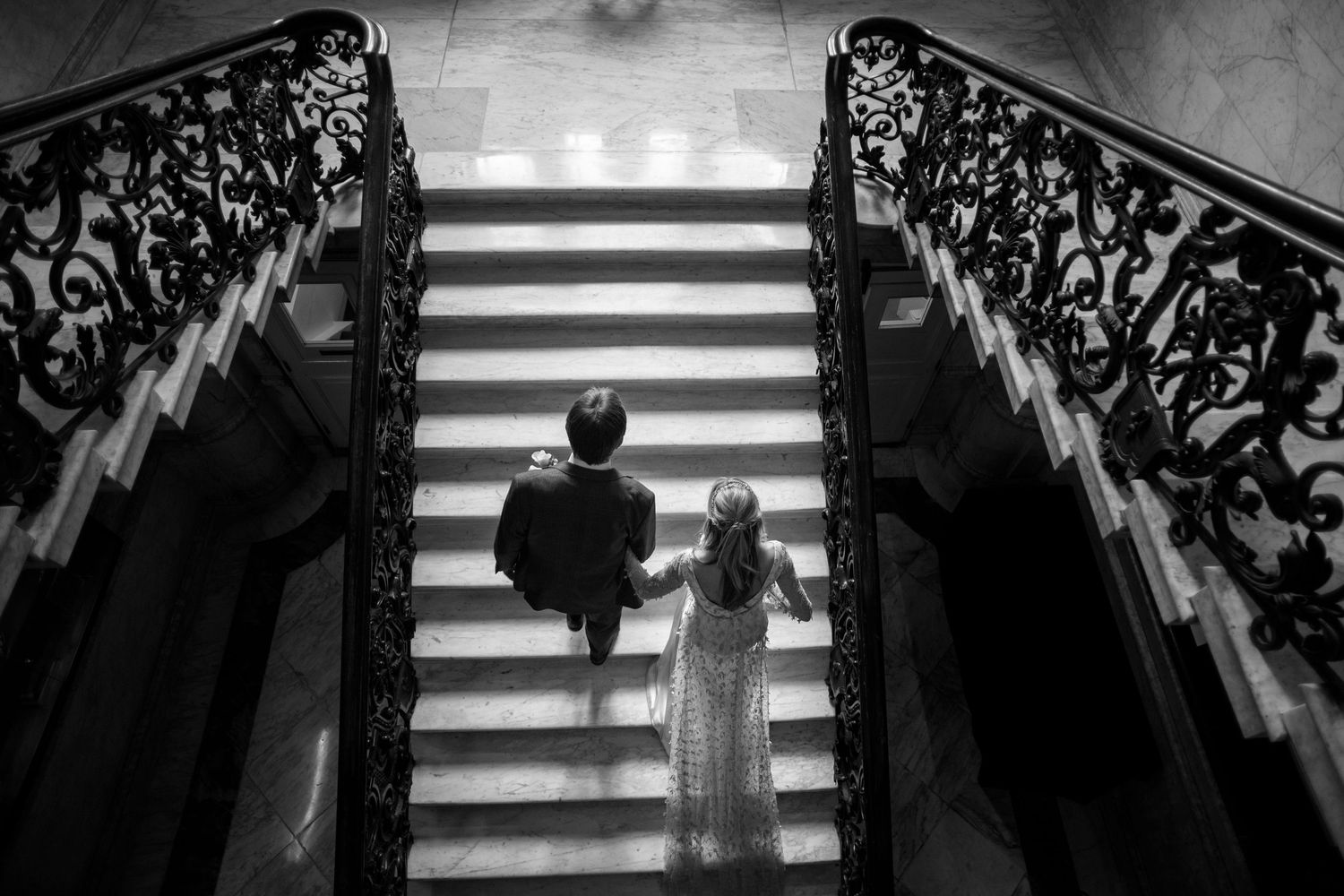 Black and white photo of bride and groom ascending ornate staircase with decorative iron railings.