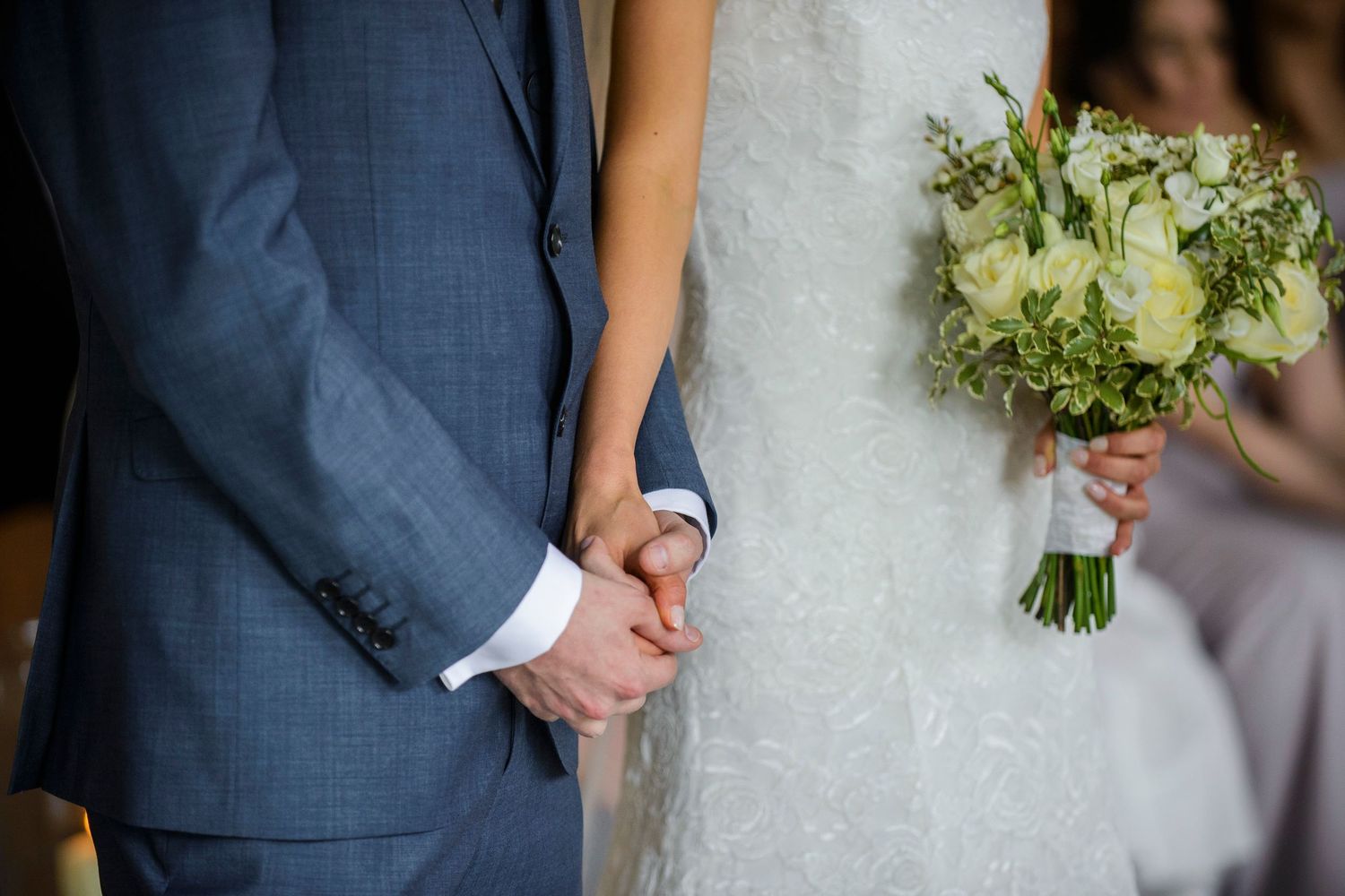 Bride and groom holding hands at wedding ceremony, bride holding white rose bouquet, groom in blue suit.