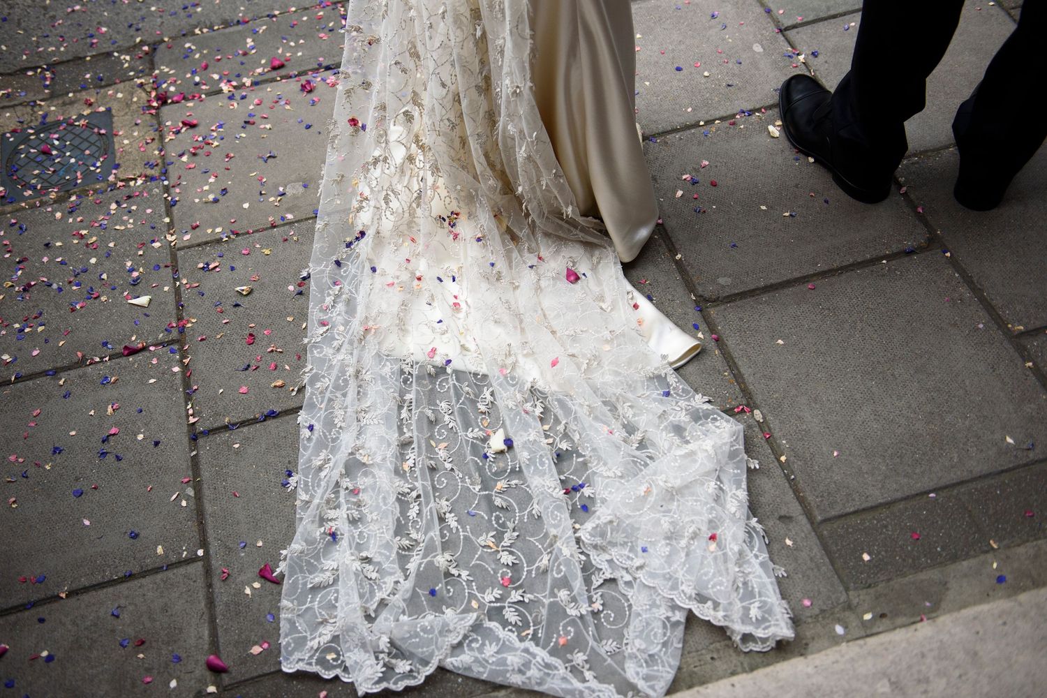 Close-up of a lace wedding dress train scattered with colorful confetti on a stone pavement.