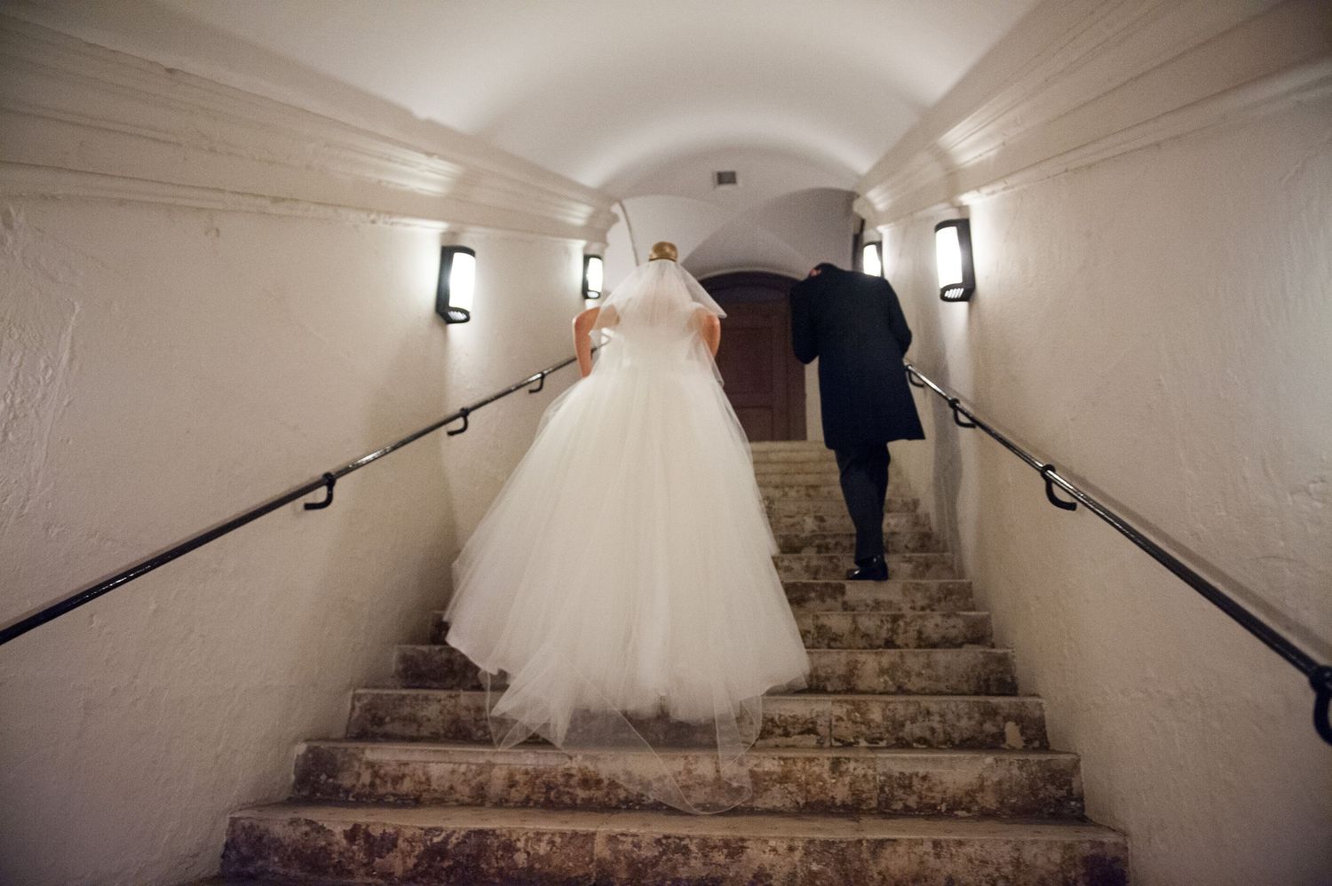 Bride and groom walking up stone stairs in a vaulted corridor, viewed from behind, wedding dress trailing.