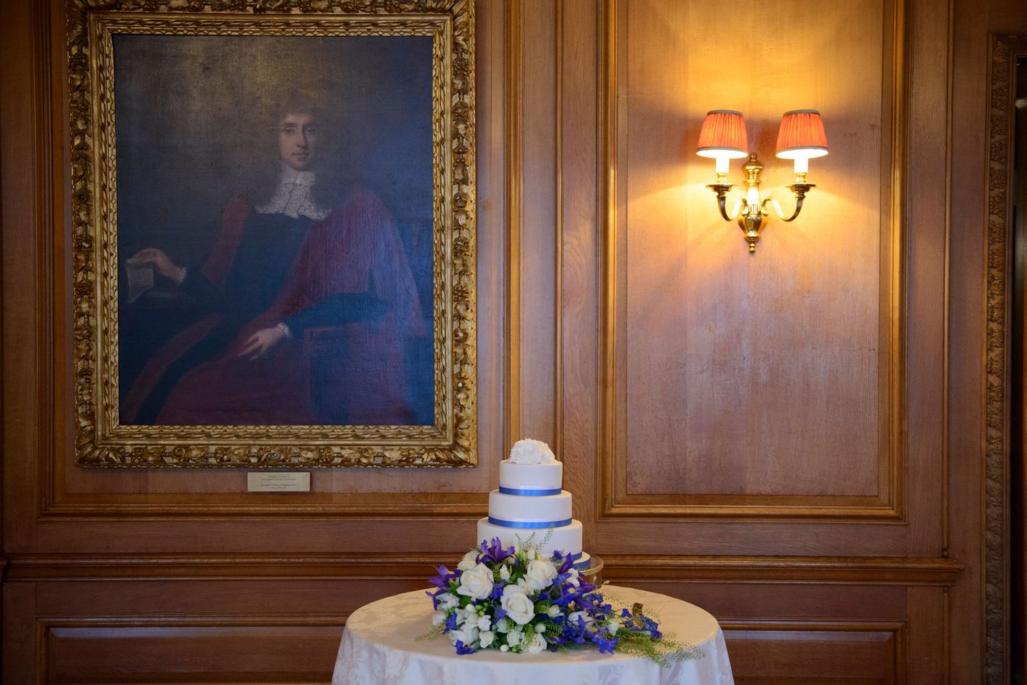 Elegant wedding cake with floral arrangement on table beneath portrait painting and wall sconce in wood-paneled room.