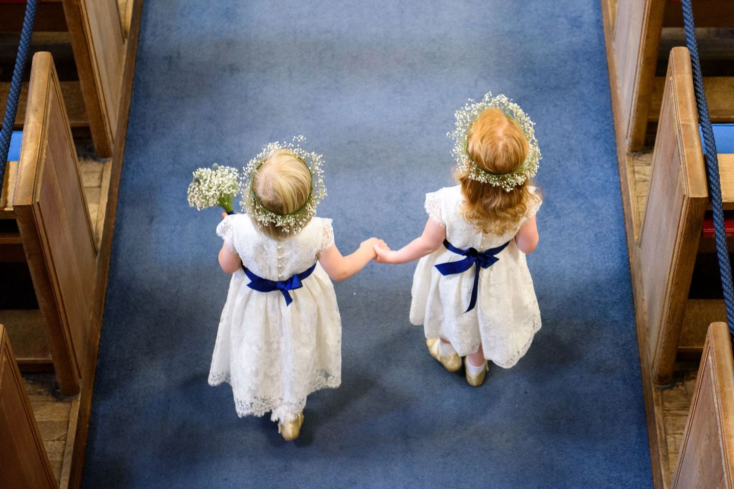 Two flower girls in white dresses holding hands walking down a church aisle from above.
