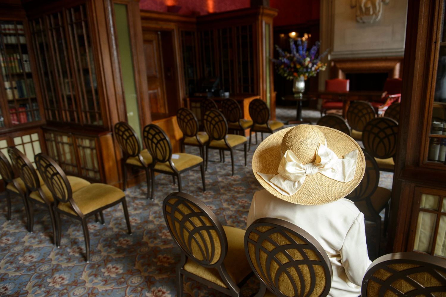 Elegant room with wooden chairs arranged in rows, a person wearing a wide-brim straw hat seated near a fireplace.