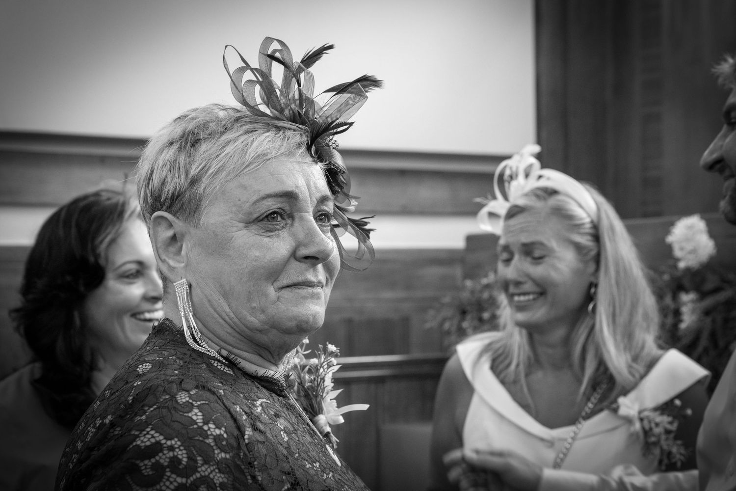 Black and white photo of elegant women at a wedding, featuring a fascinator hat with feathers.