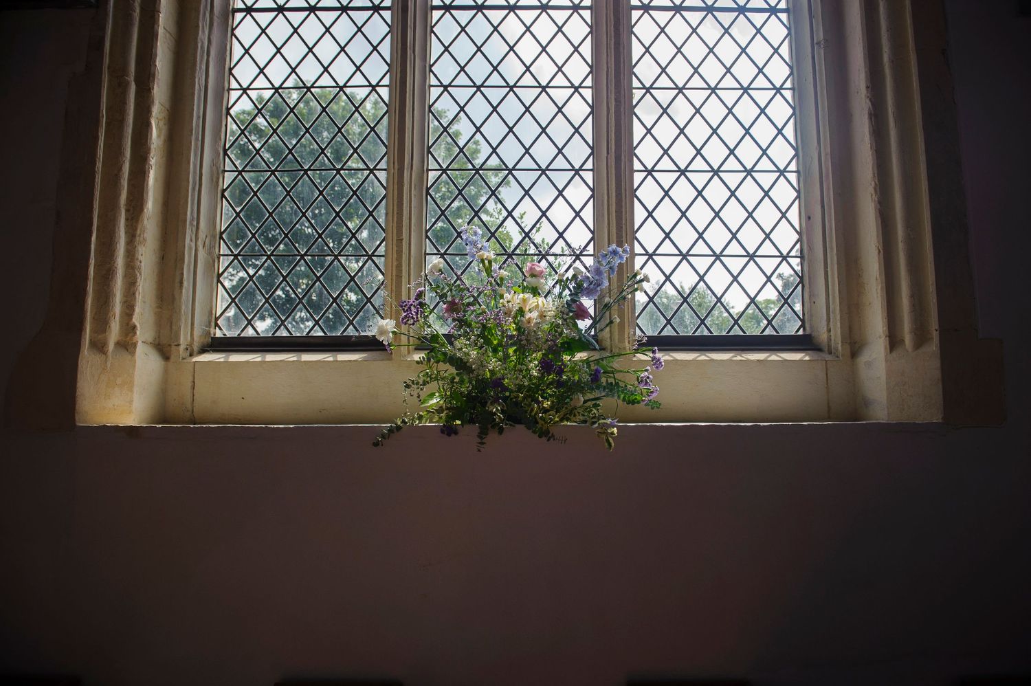 Floral arrangement on a church windowsill with leaded glass window and green garden view beyond.