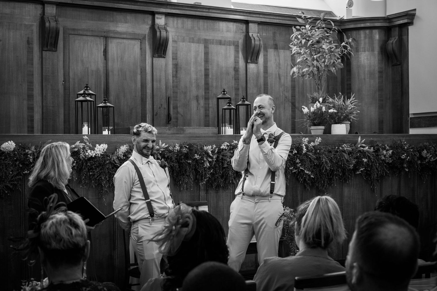 Two grooms share vows at their wedding ceremony before seated guests in a decorated hall.