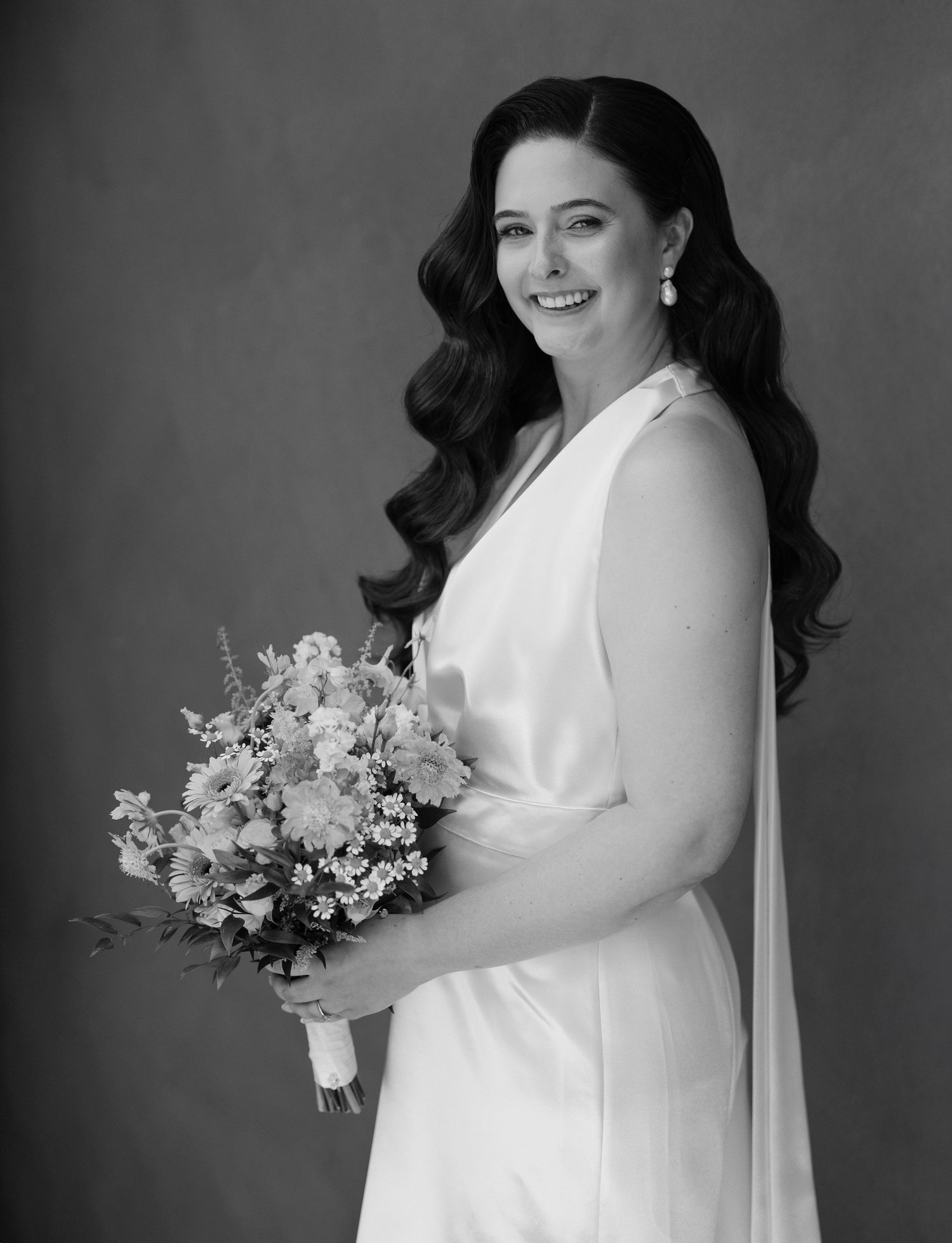 Elegant bride with long dark wavy hair holding a wildflower bouquet in a white gown, black and white portrait.
