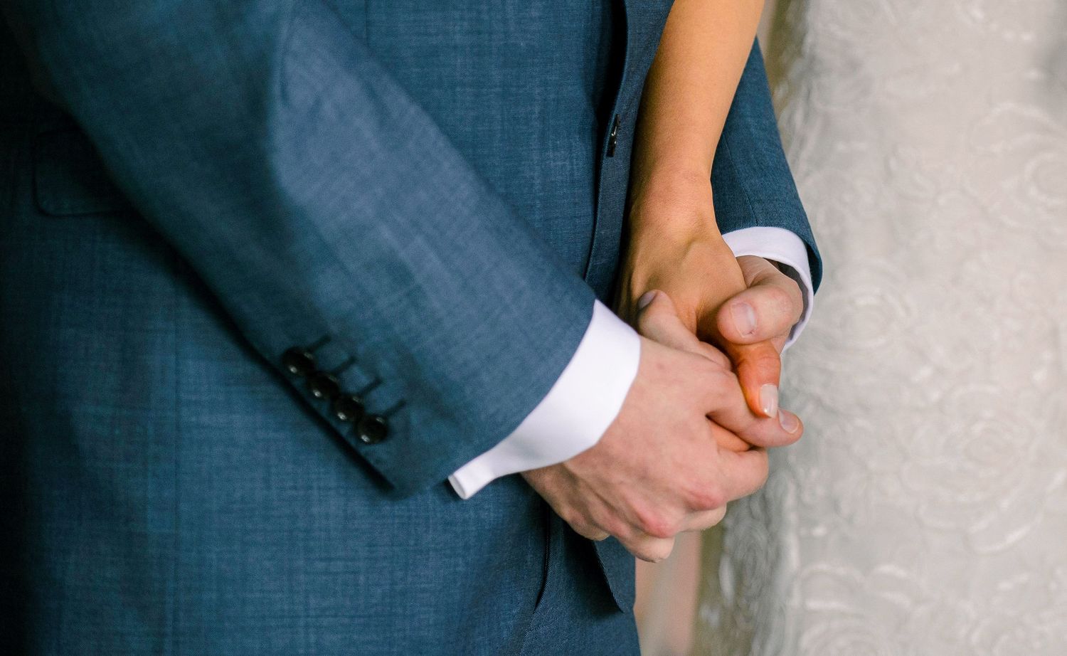Close-up of bride and groom holding hands, groom in blue suit with white shirt cuff visible.