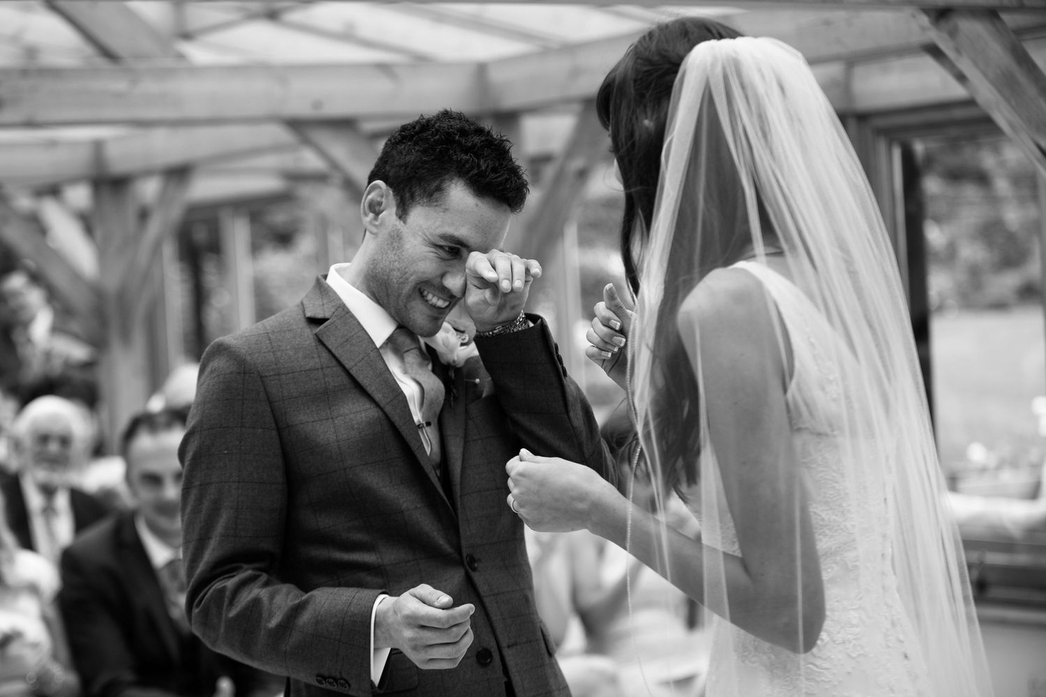 Emotional groom wiping tears as bride approaches during outdoor wedding ceremony in black and white photo.