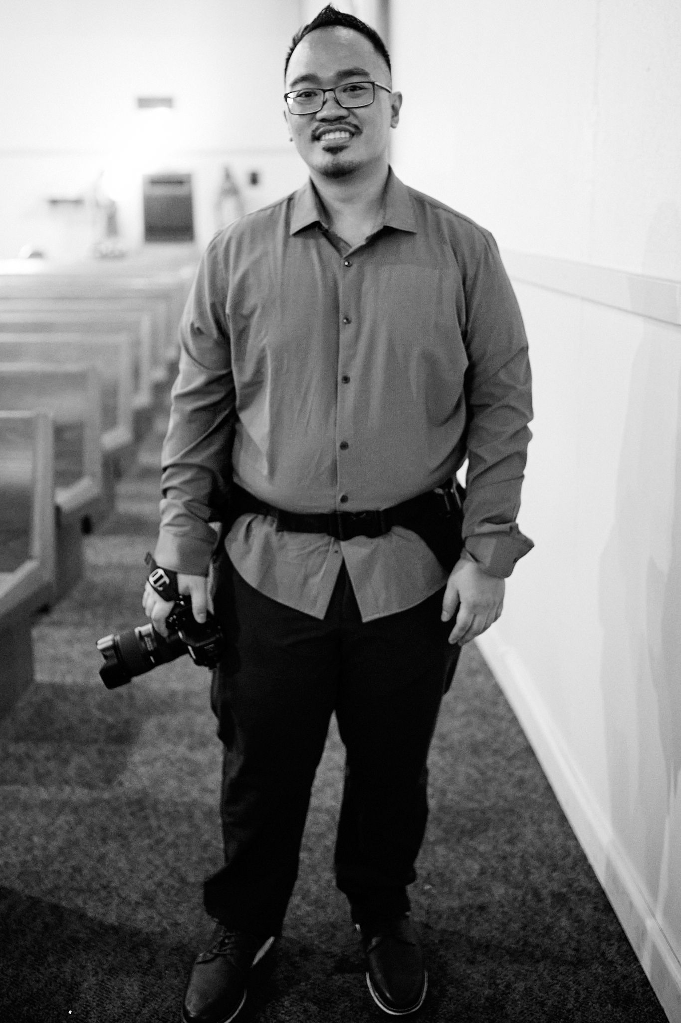 A photographer in a gray shirt and black pants poses with a camera in a black and white portrait series.