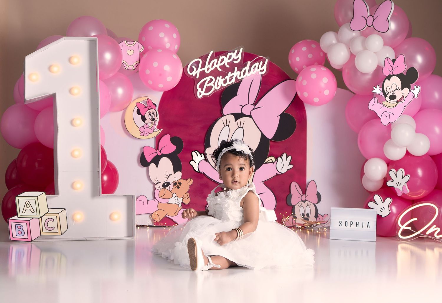 Baby girl in white dress at Minnie Mouse first birthday party with pink balloons and decorative backdrop.