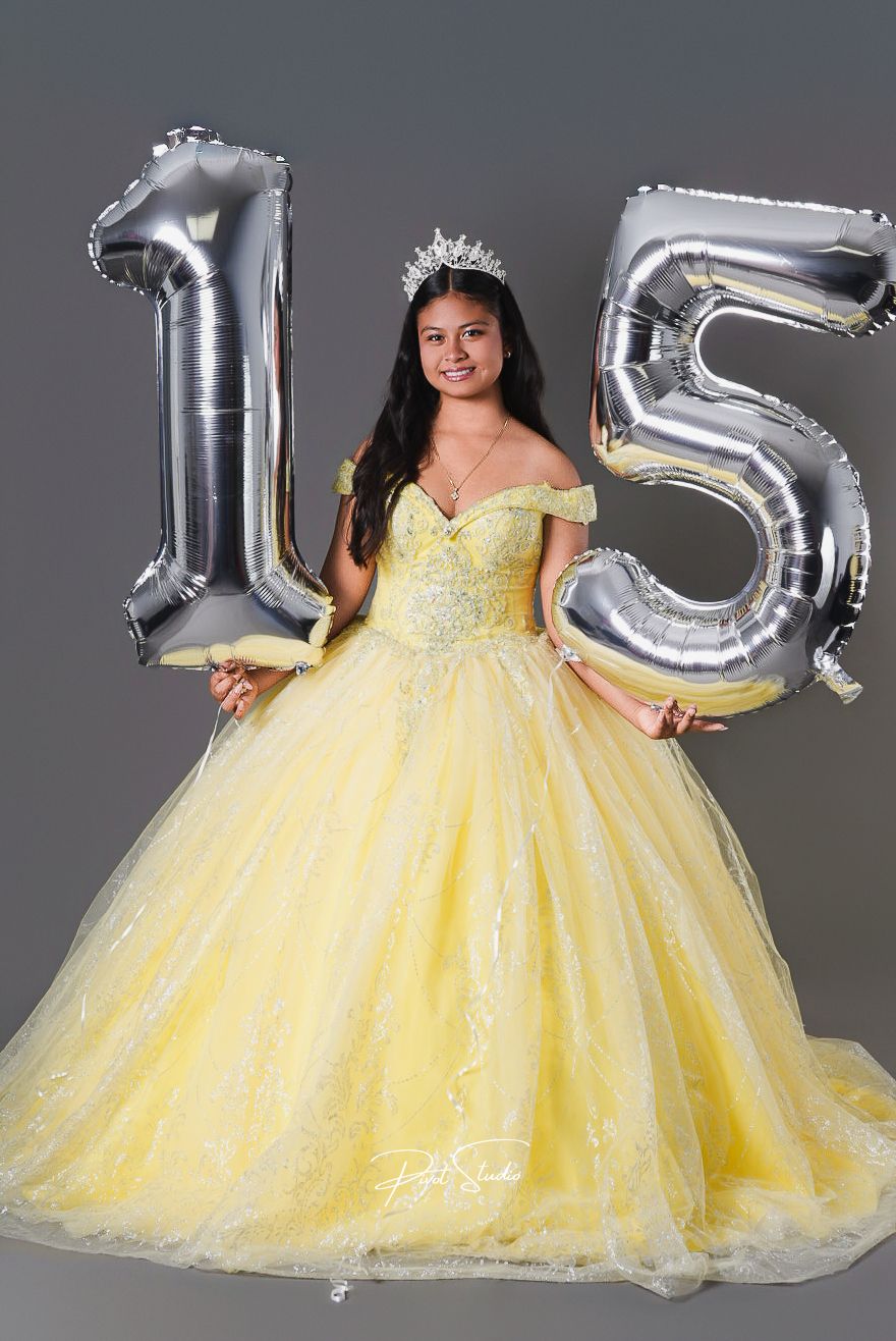 Young woman in yellow ball gown with tiara holds silver number 15 balloons for her Quinceanera celebration.