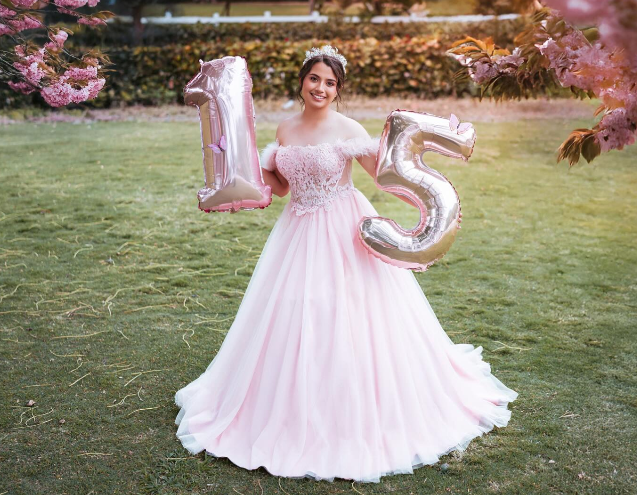 Young woman in pink ball gown holding silver number 15 balloons outdoors for quinceañera celebration.