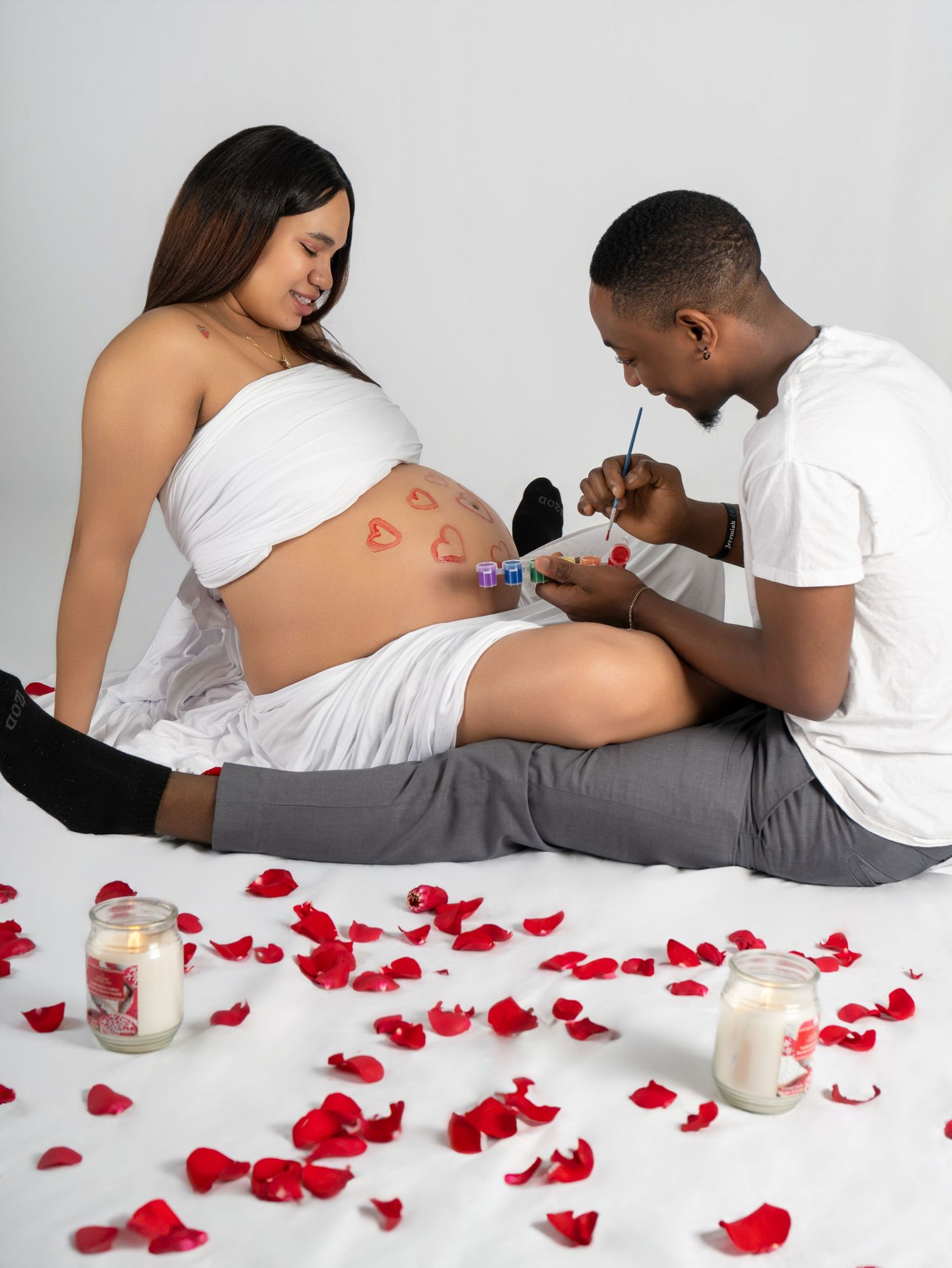 A man paints hearts on a pregnant woman's belly surrounded by red rose petals and candles in a romantic maternity photo shoot.