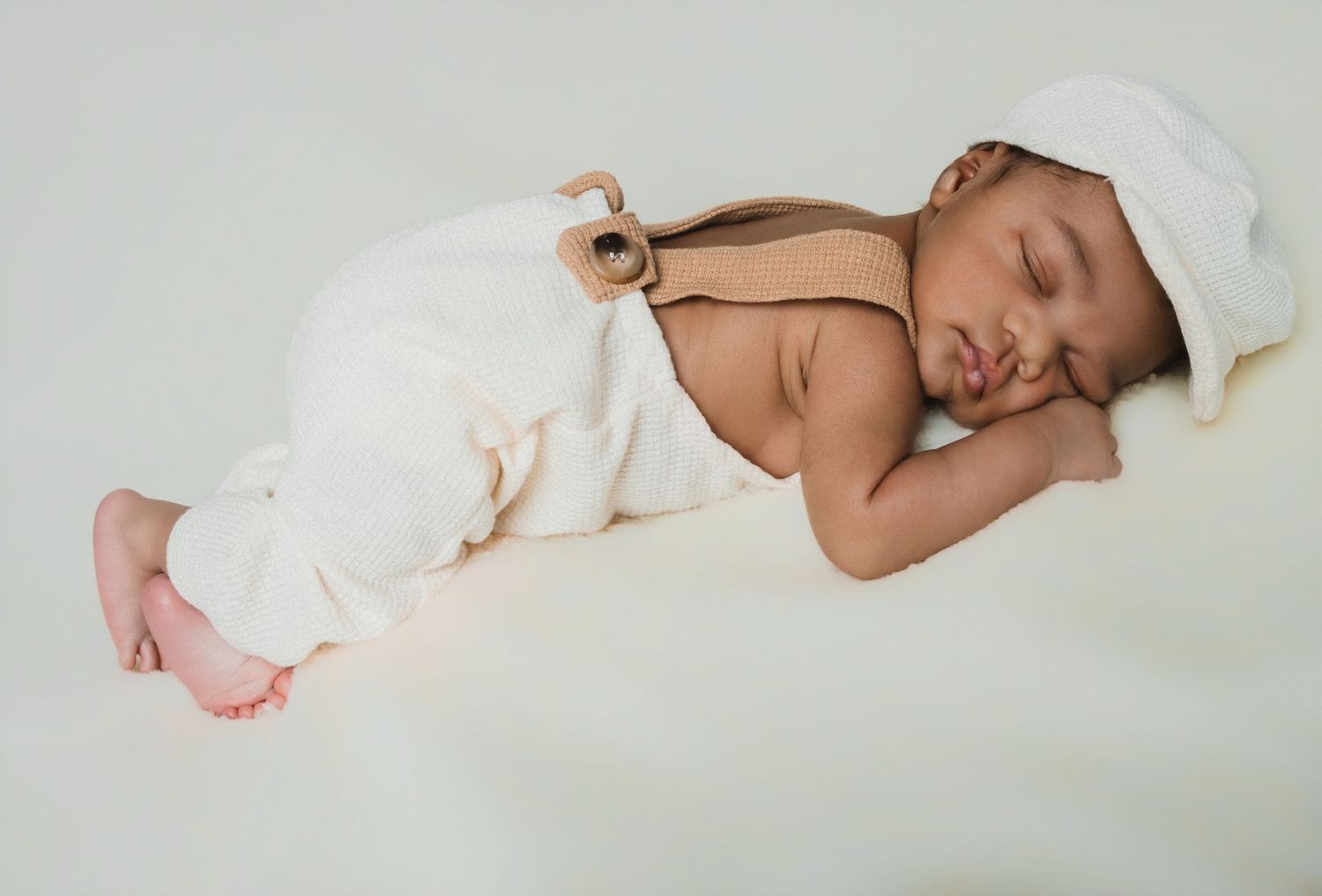 Newborn baby sleeping peacefully in white overalls and hat on a soft white background.