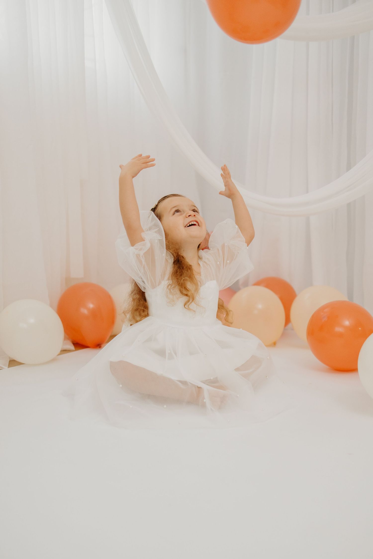 Child in white dress reaching up towards orange and white balloons against draped fabric backdrop.