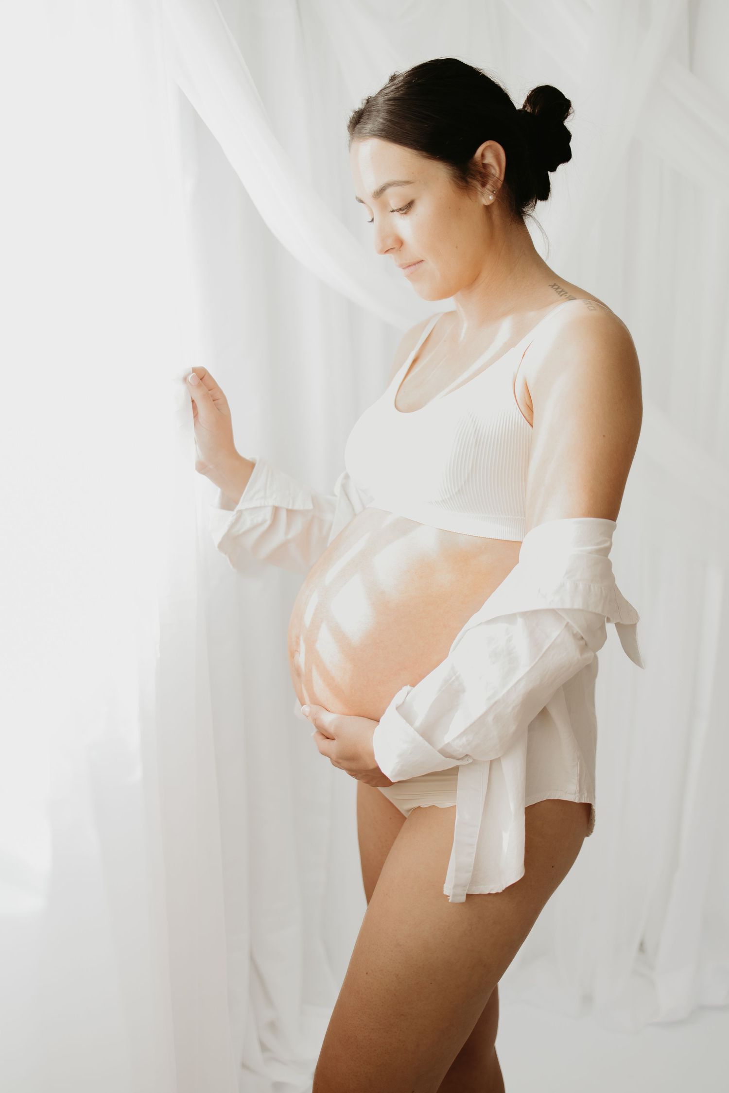 Expecting mother in white lingerie poses gently against light curtains in a soft maternity photoshoot.