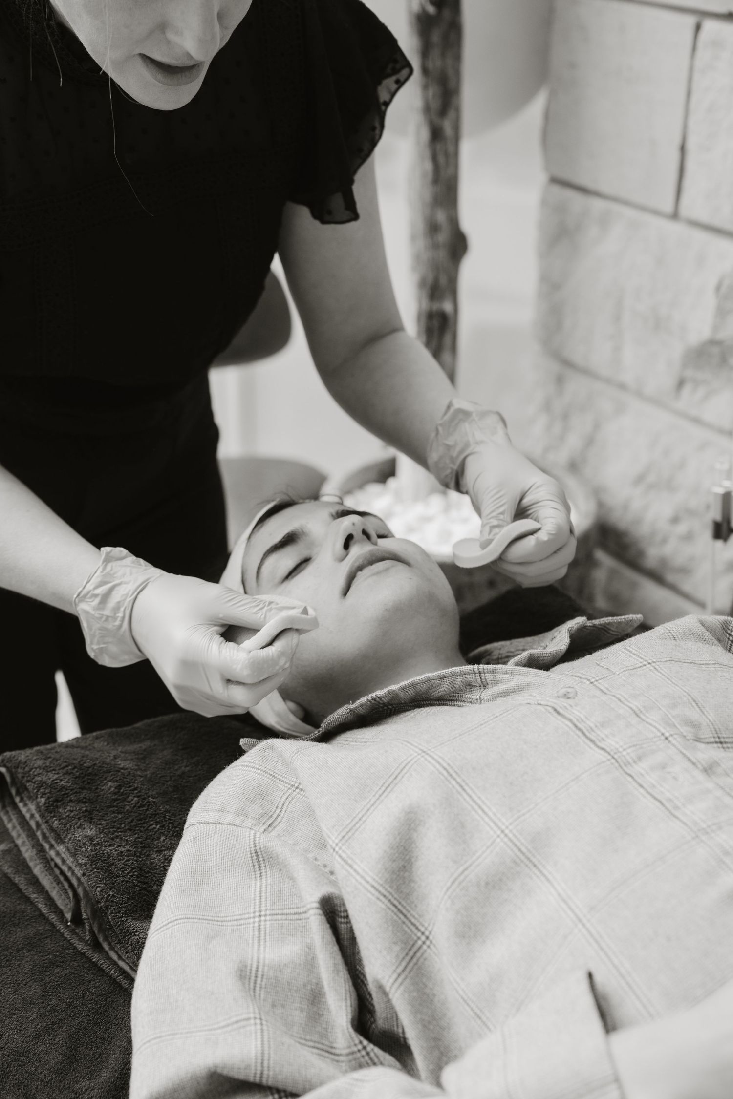 A black and white image shows someone receiving a facial treatment or massage while lying on a table, with gloved hands working.