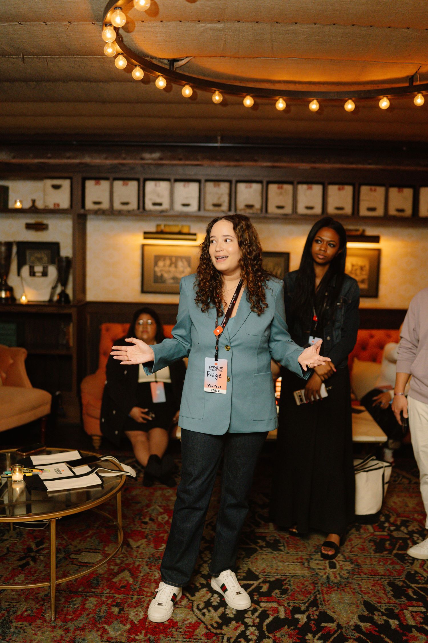 A speaker gestures while presenting in a dimly lit venue with string lights and patterned carpet.