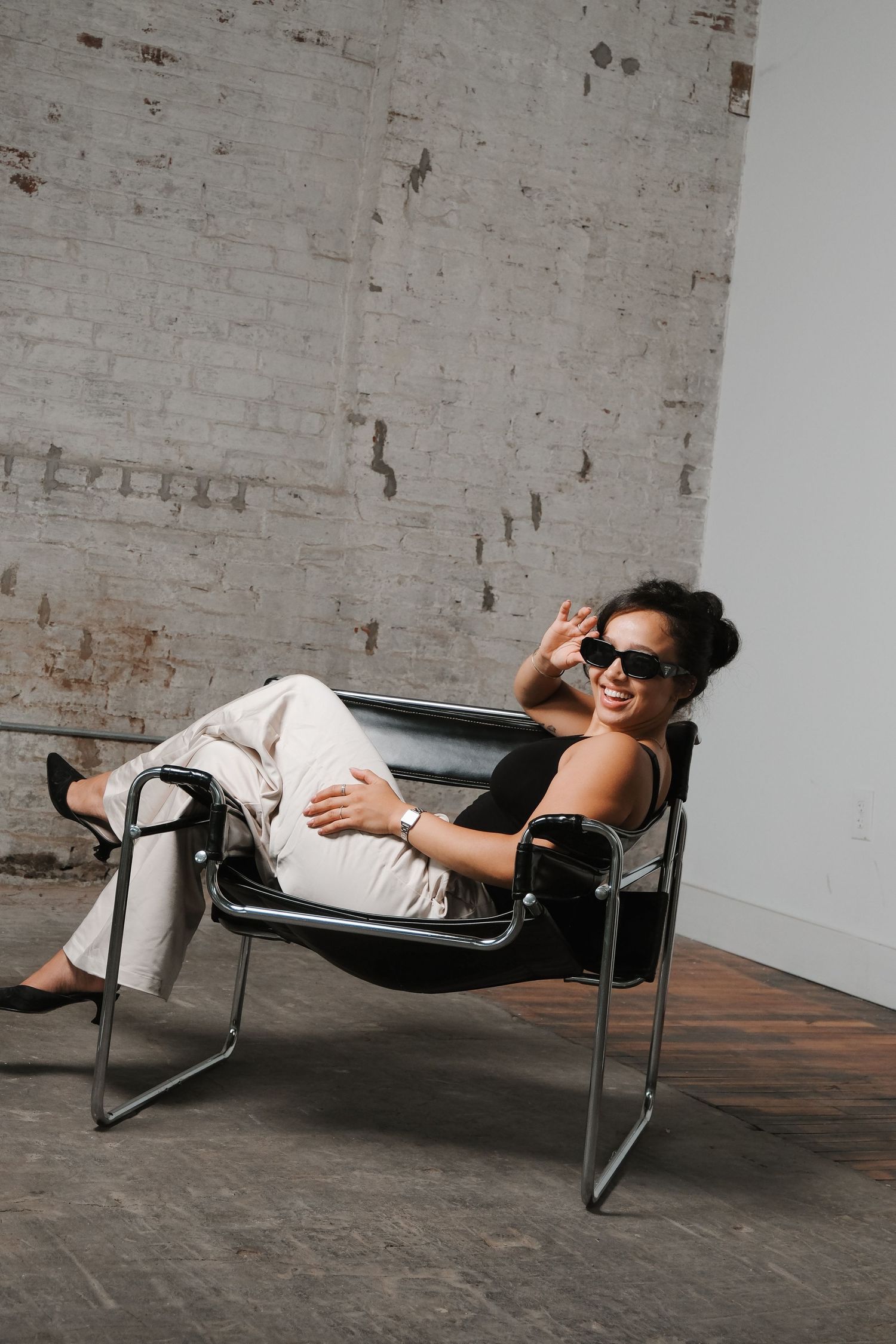A person in sunglasses relaxes casually on a chrome and leather Wassily chair against an industrial-style brick wall.