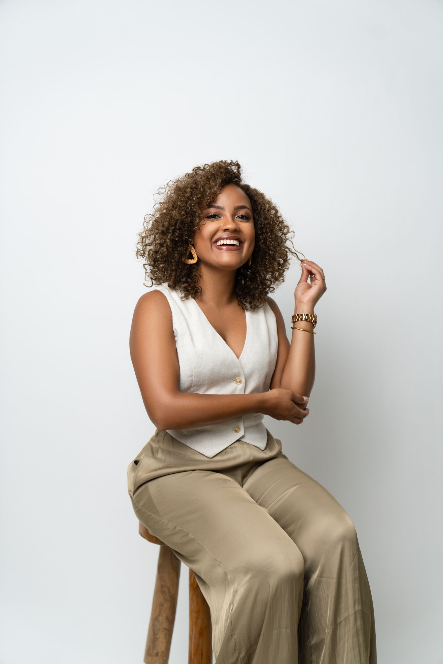 A woman wearing a white sleeveless top and beige wide-leg pants sits casually on a wooden stool against a white background.