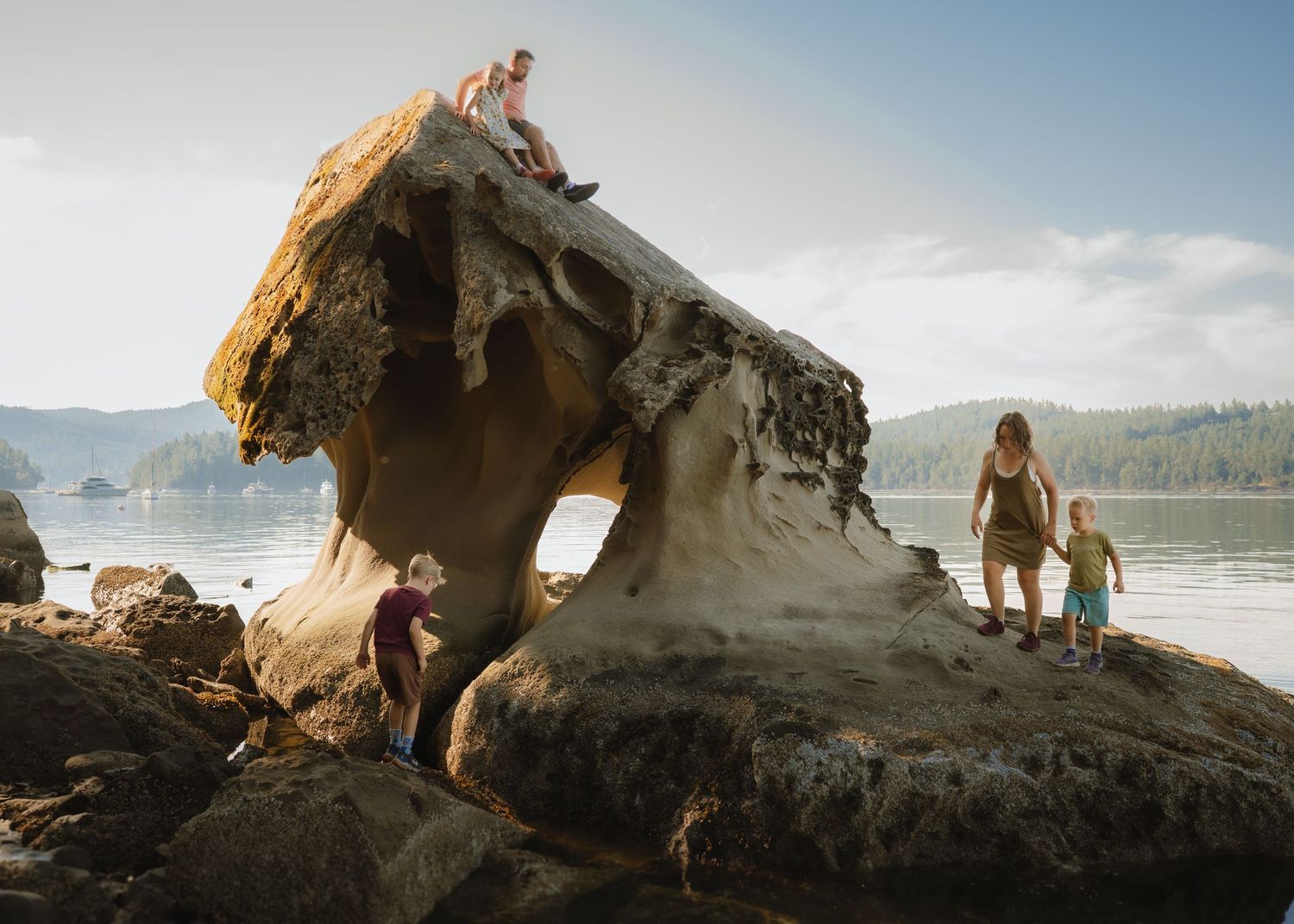 Adults and children explore a large sandstone rock sculpted by erosion on a quiet coastal beach with forested hills beyond.