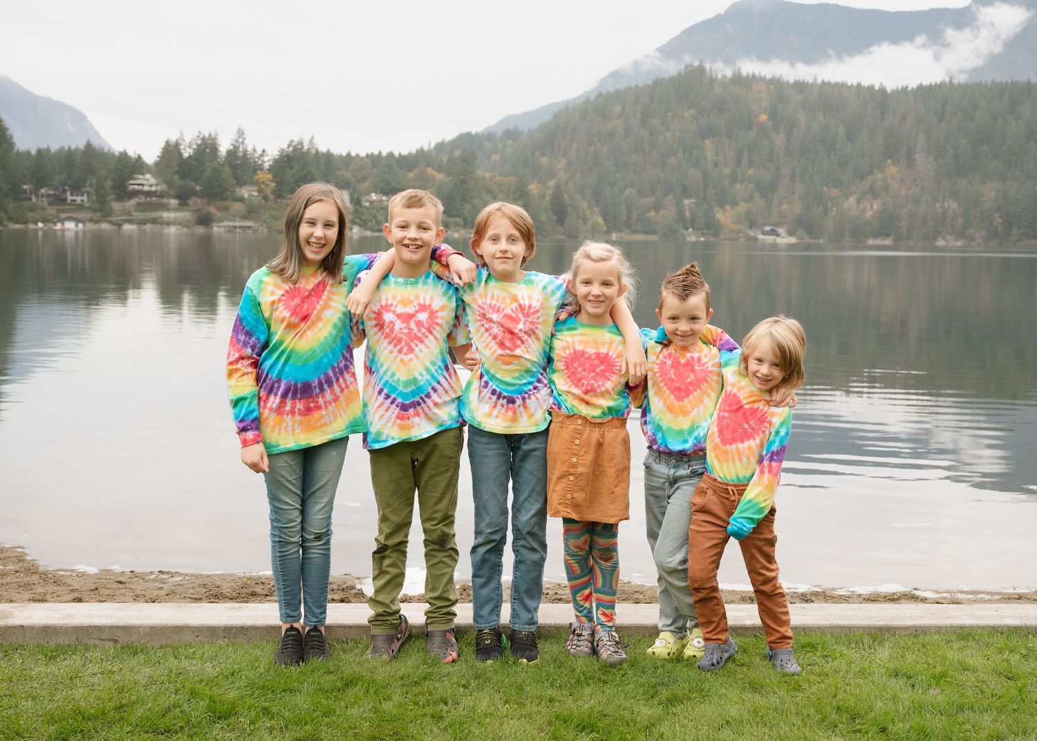 Seven children in colorful tie-dye shirts pose with arms around each other on a lakeside lawn with forested mountains behind.