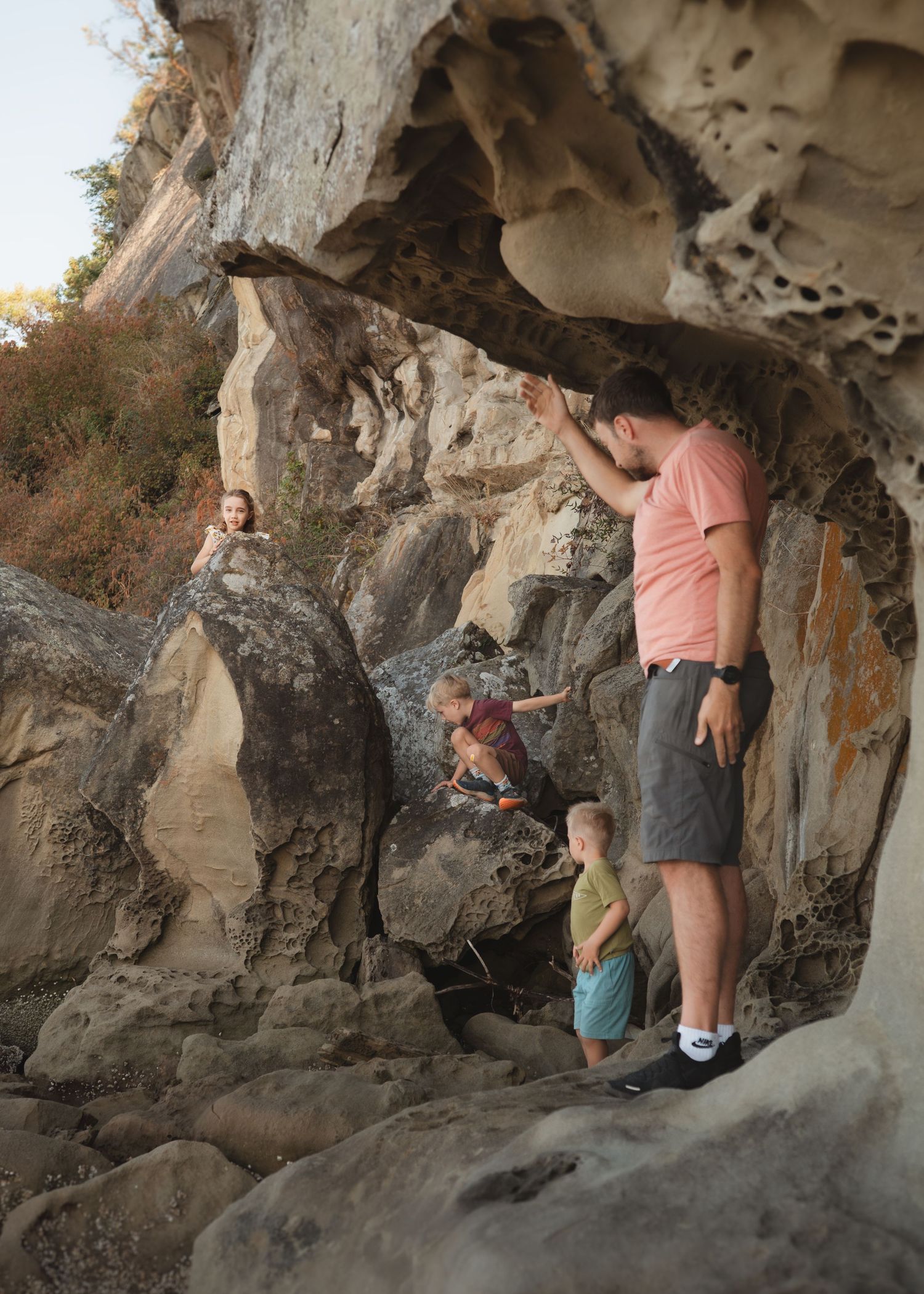 A man in a pink shirt and shorts points up at a sandstone rock overhang while three children explore the boulders below.