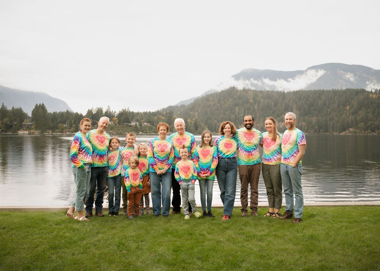 Large family in matching rainbow tie-dye shirts stands together on a grassy lakeshore with forested mountains in the background.