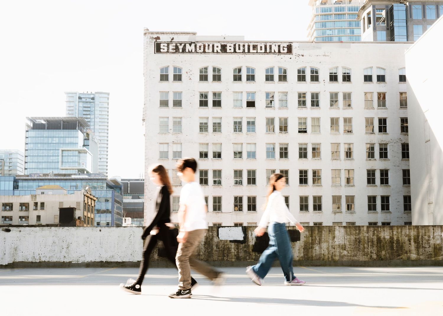 Three people walk past the Raymond Building exterior in an urban setting, with sunlight casting long shadows.