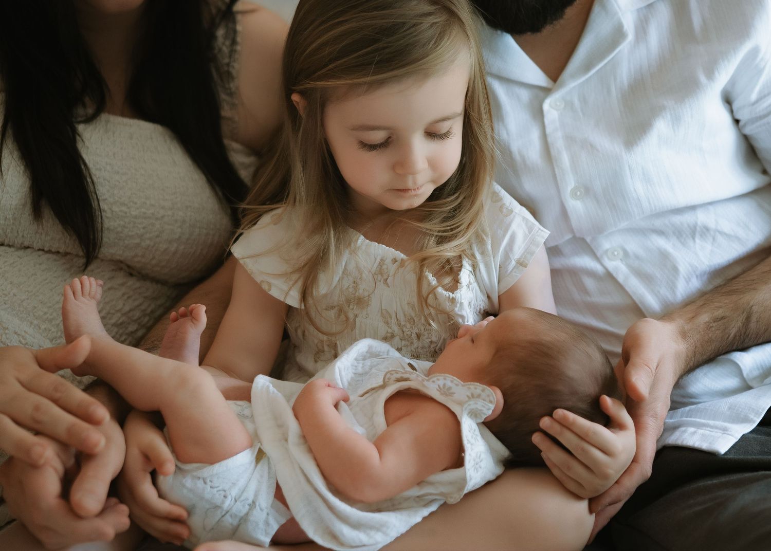 A toddler girl gazes down at her newborn sibling resting on her parents' laps in an intimate family newborn portrait.