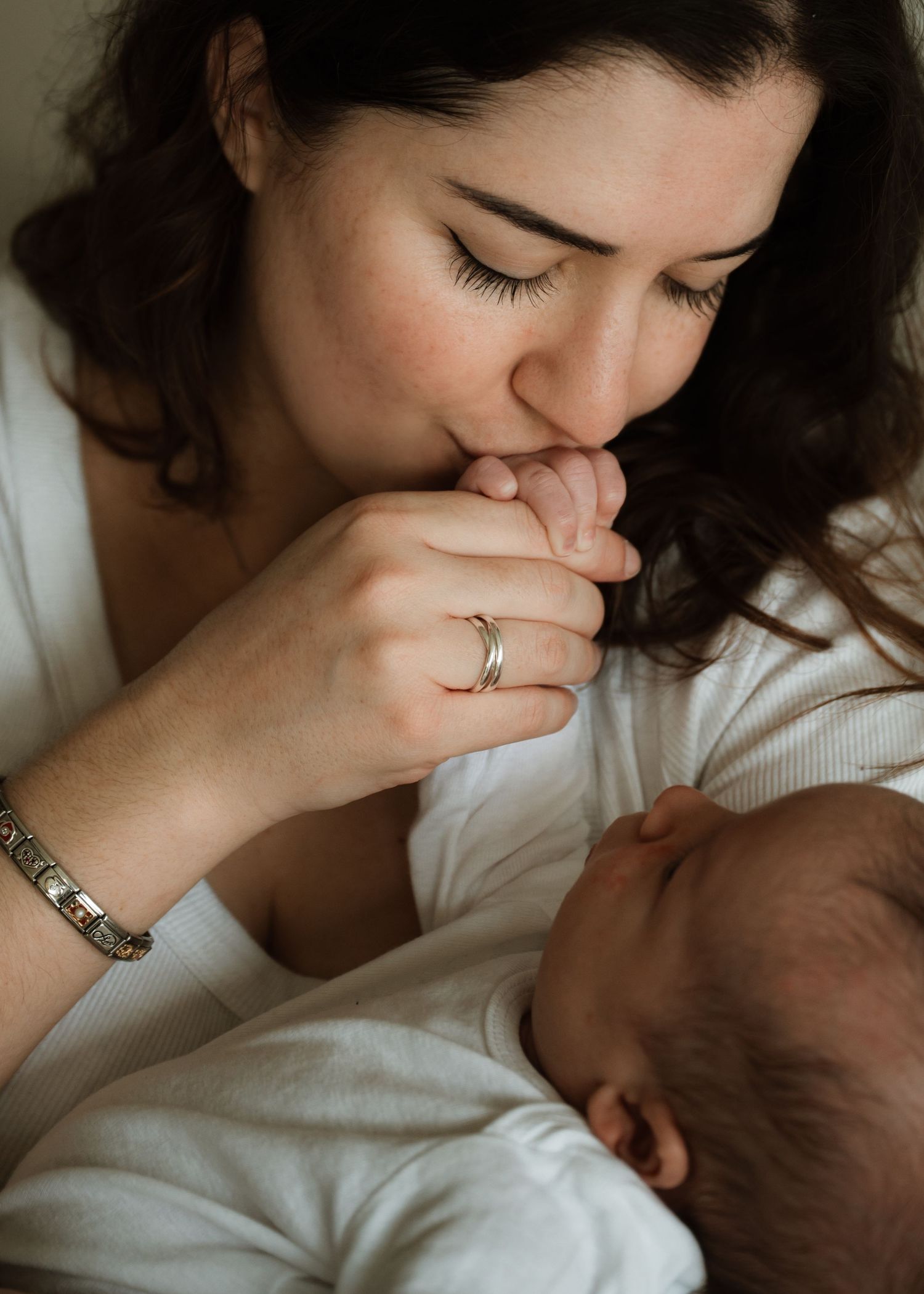 Mother tenderly kisses her newborn baby's tiny hand while holding the sleeping infant close in a quiet, loving moment.