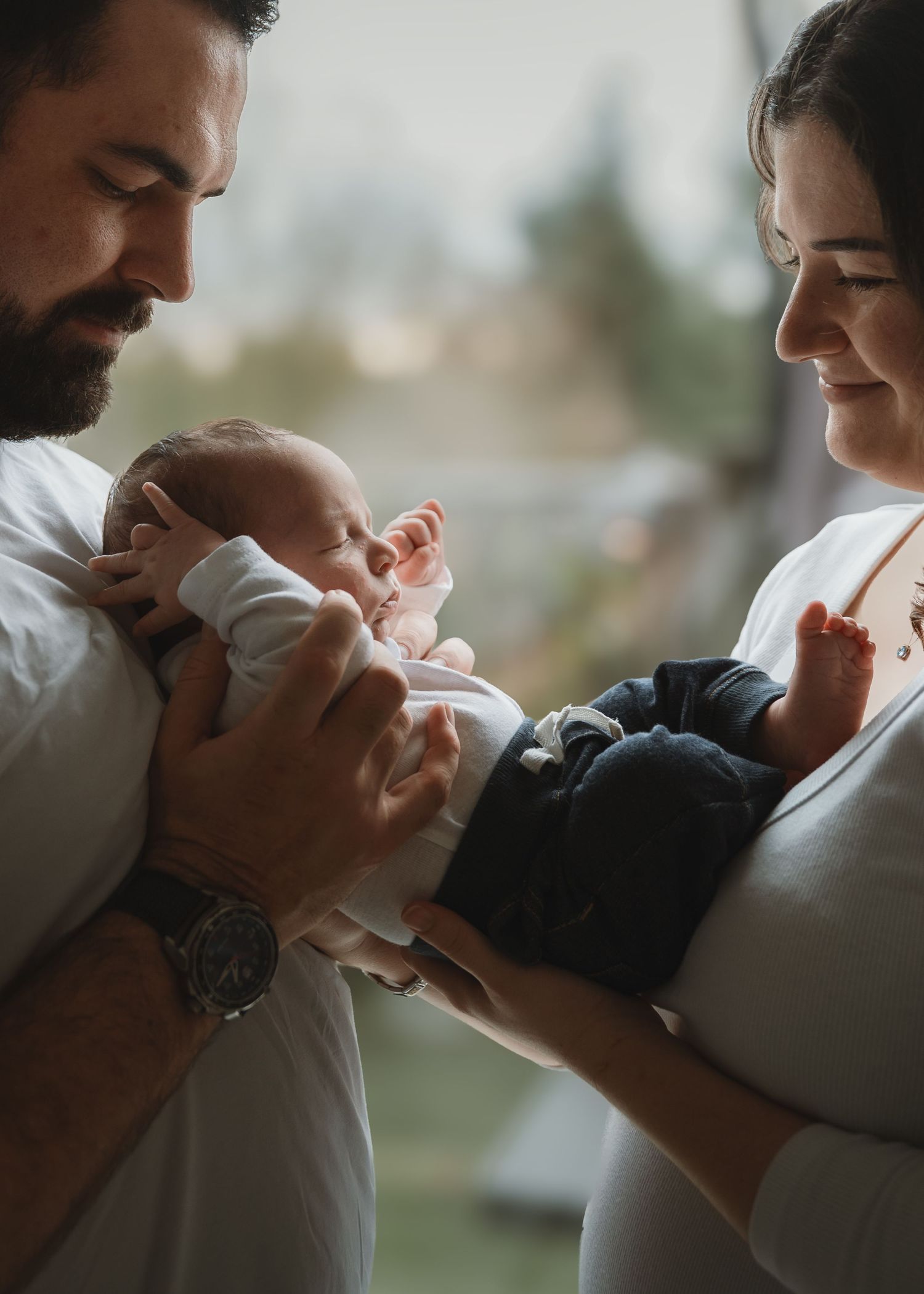 Parents hold their newborn baby between them in soft window light, looking down at the infant with tender smiles.